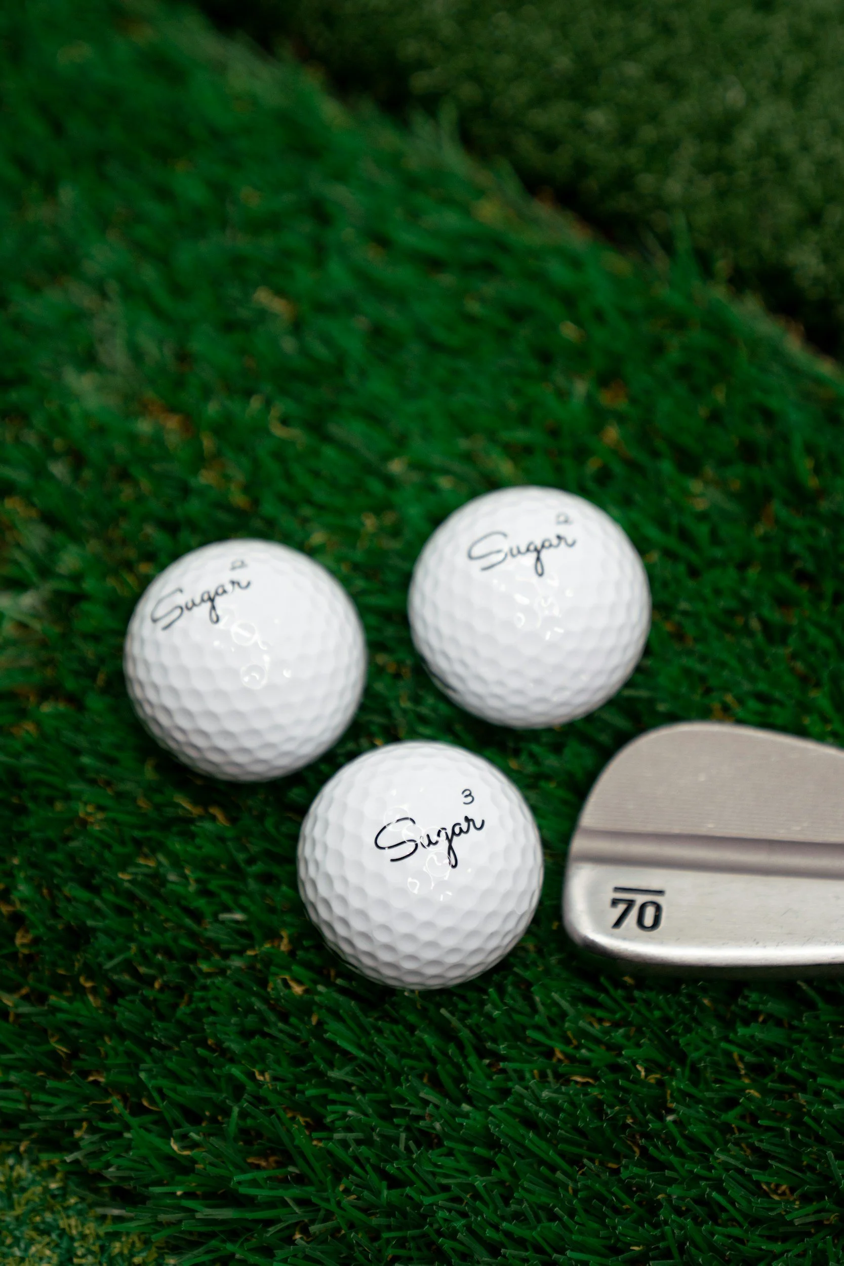 Three white golf balls marked with 'Sugar' and a golf club lying on green artificial grass.