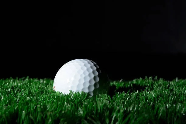 A close-up of a golf ball resting on green artificial turf under low light.