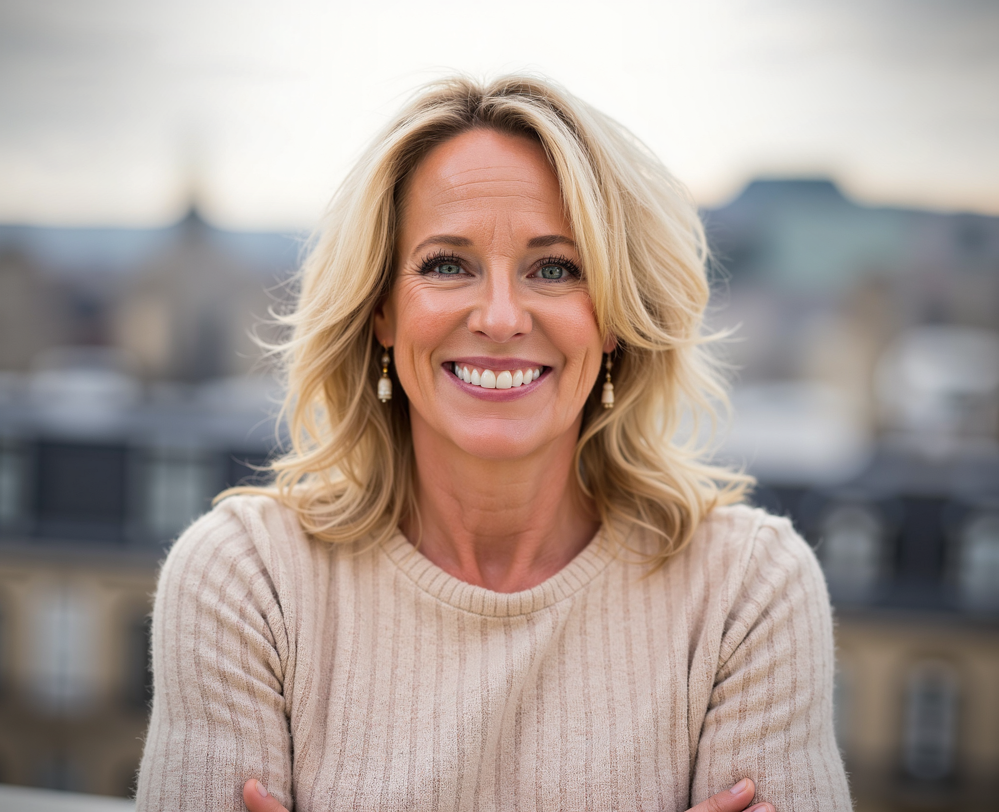 A smiling woman with blonde hair wearing a beige sweater and earrings, standing outdoors with a cityscape in the background.