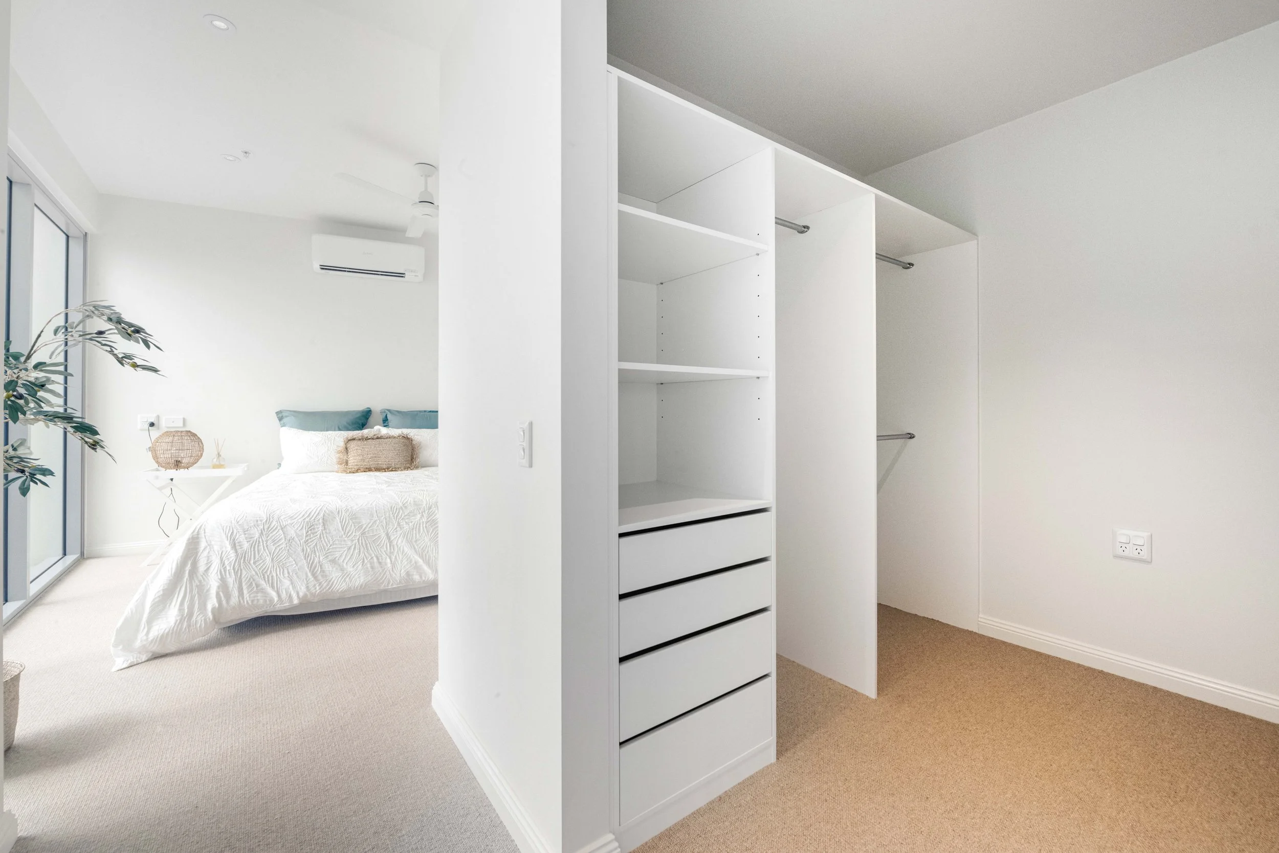 Minimalist bedroom with a double bed with white bedding and teal pillows, adjacent to a walk-in closet with white shelves and drawers.