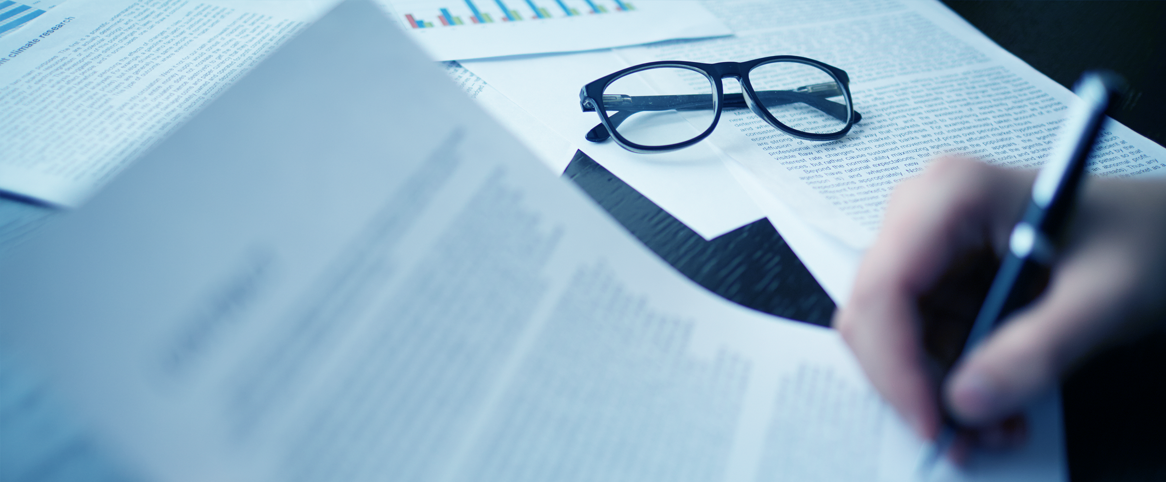 Close-up of black eyeglasses on a table with scattered papers, hand writing with a pen.