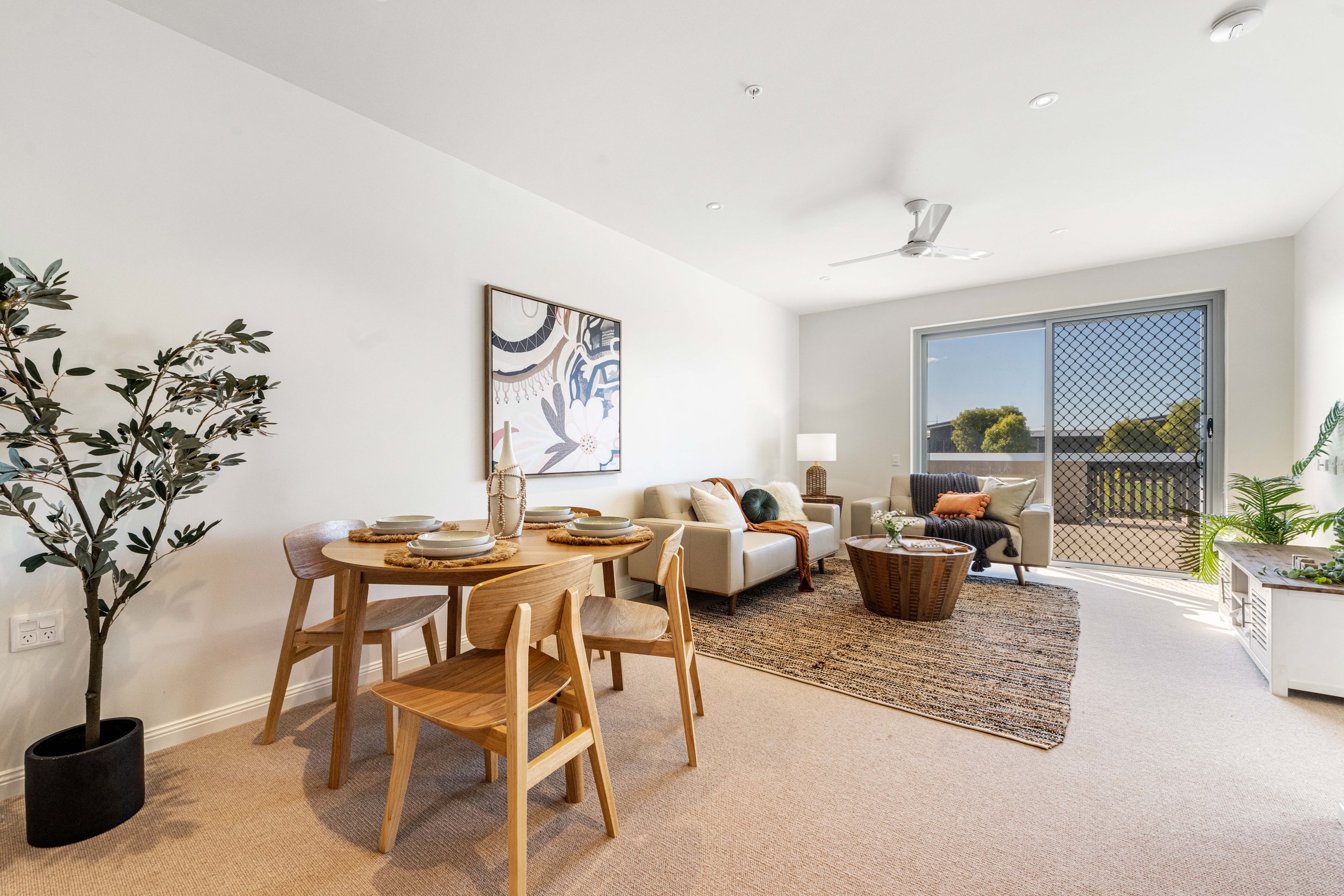 Modern living and dining area with a small wooden table set for four, beige sofa, accent chair, indoor plants, wall art, and sliding door to a balcony.