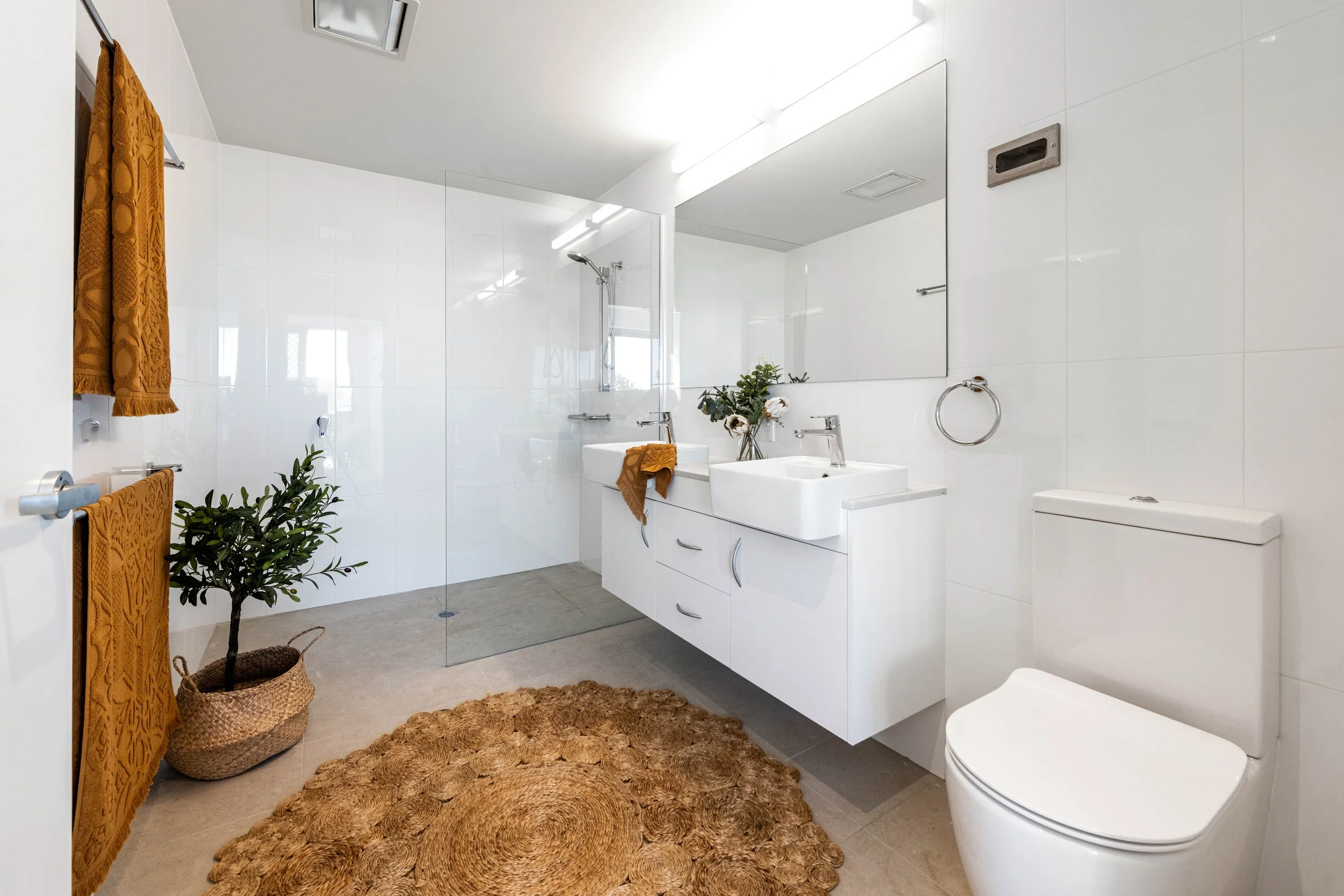 Modern bathroom with white fixtures, dual sinks, large mirror, and glass shower. Features orange towels, woven basket with plant, and circular woven rug. Minimalist design with clean, bright aesthetic.