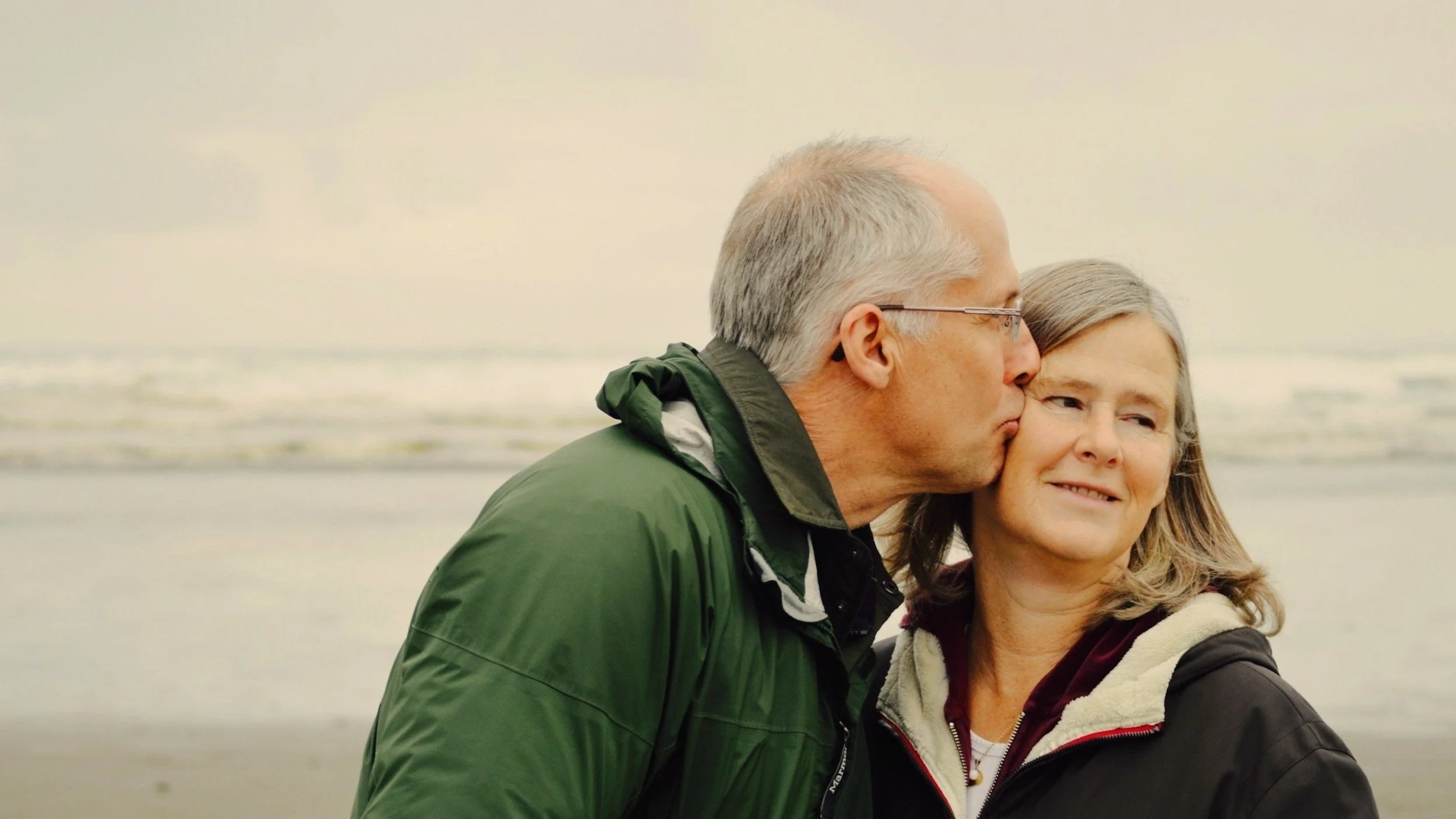Older man kissing older woman's cheek at the beach.