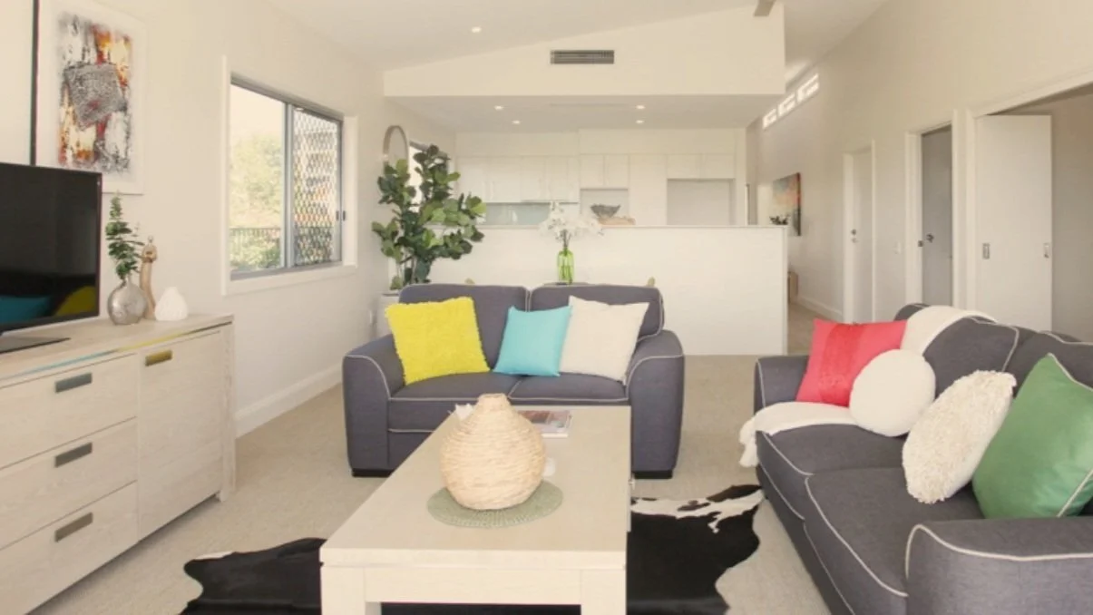 Modern living room with gray sofas, colorful pillows, a coffee table, TV stand, and wall art; open kitchen in the background and a large window with outdoor view.