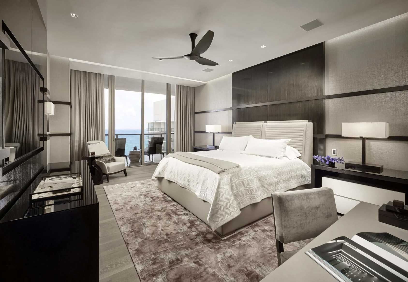 Modern hotel bedroom with a large white bed, a balcony with ocean view, and contemporary furniture.