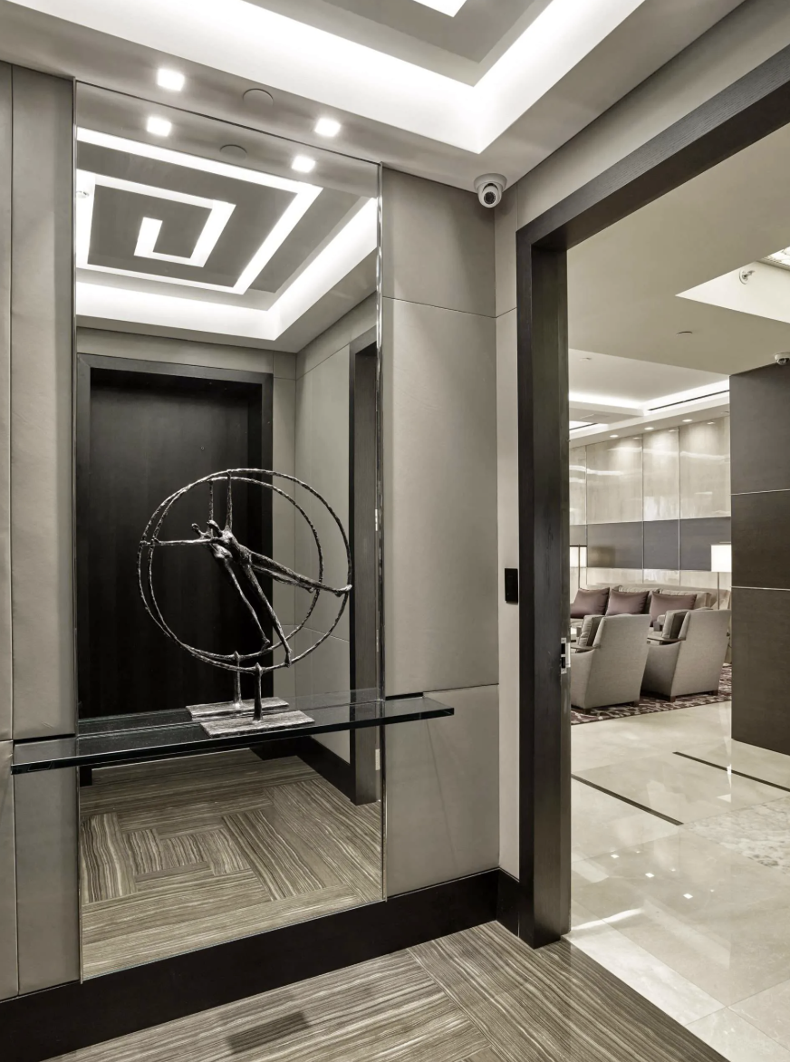 Modern hotel lobby with artwork sculpture on glass-topped table, mirror reflecting ceiling lights and geometric ceiling design, beige and brown tones, comfortable chairs and reception area in background.