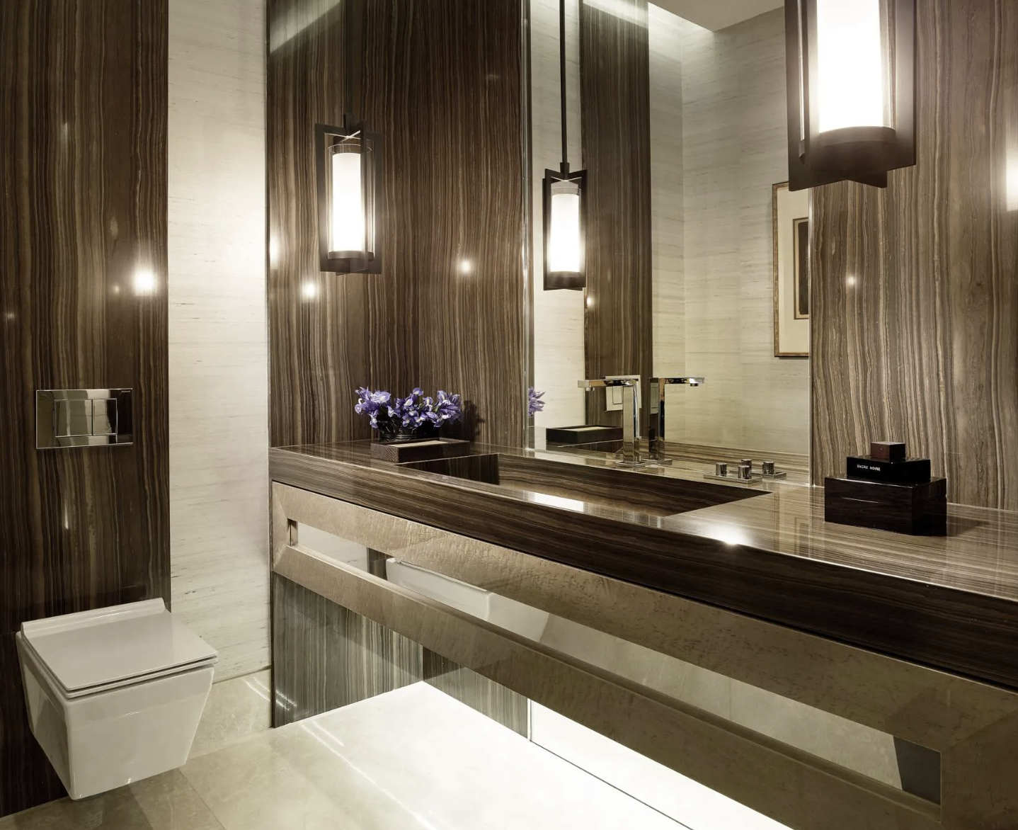 Modern bathroom with large mirror, brown and beige marble surfaces, purple flowers in a vase, and contemporary lighting fixtures.