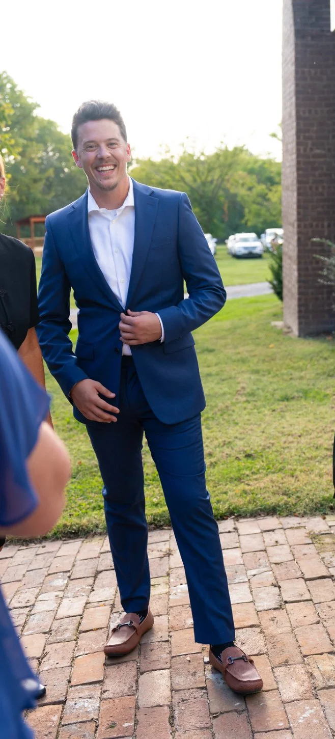 A smiling man in a blue suit and white shirt standing outdoors on a brick pathway with green grass and trees in the background.