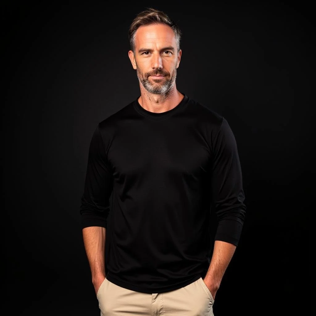 A middle-aged man with short gray hair and a beard stands against a dark background, wearing a black long-sleeve shirt and beige pants.