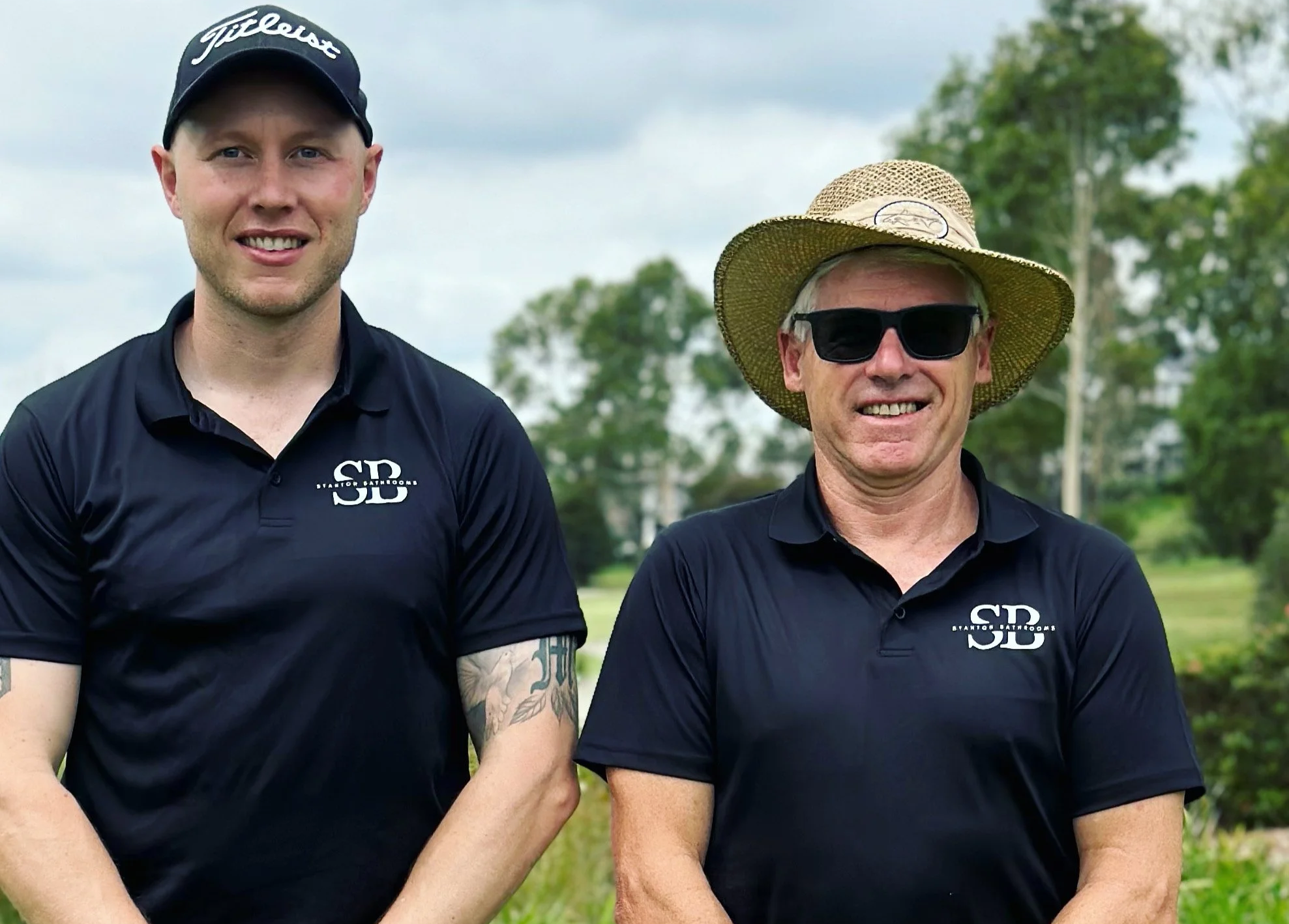 Two men wearing black polo shirts stand outdoors on grass. The man on the left wears a baseball cap, while the man on the right wears a straw hat and sunglasses. Trees and a sign are visible in the background.