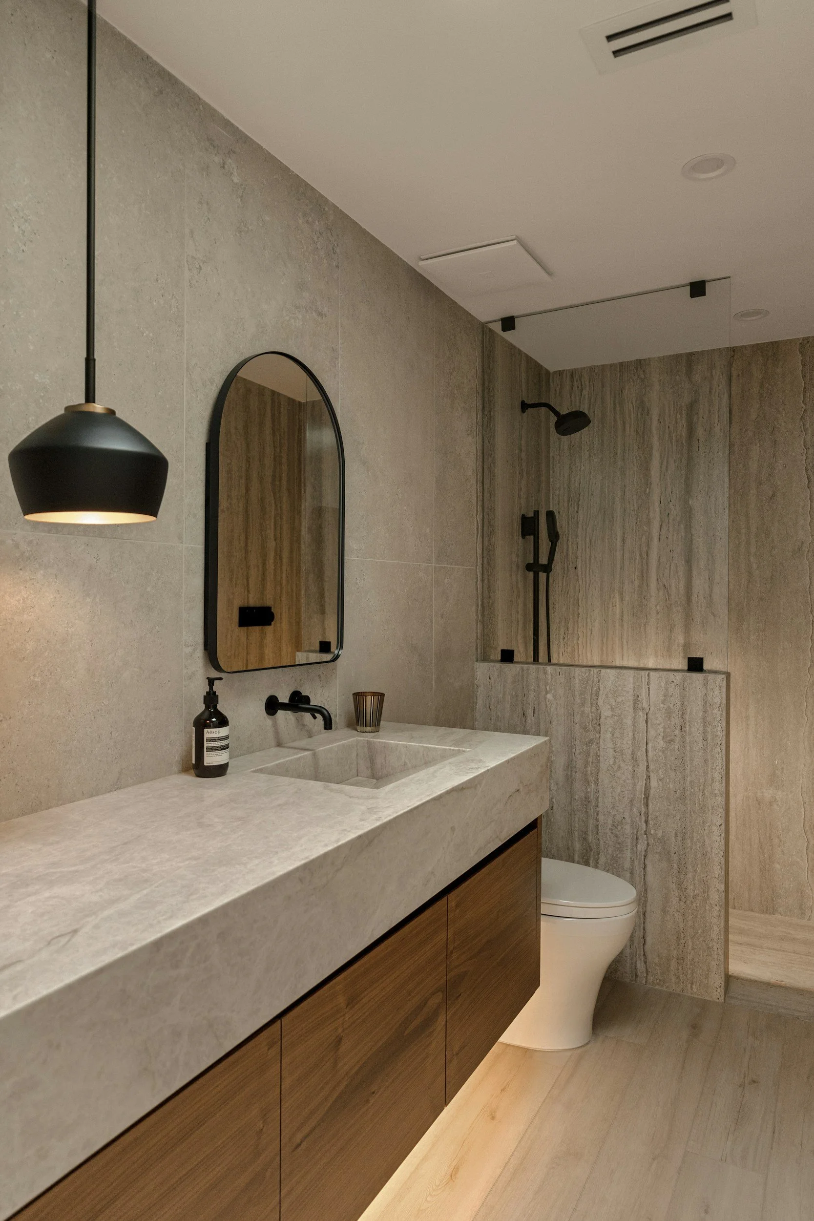 Modern bathroom with beige stone walls, wood vanity with a stone countertop, oval mirror, black fixtures, a shower with black fixtures, and a white toilet.