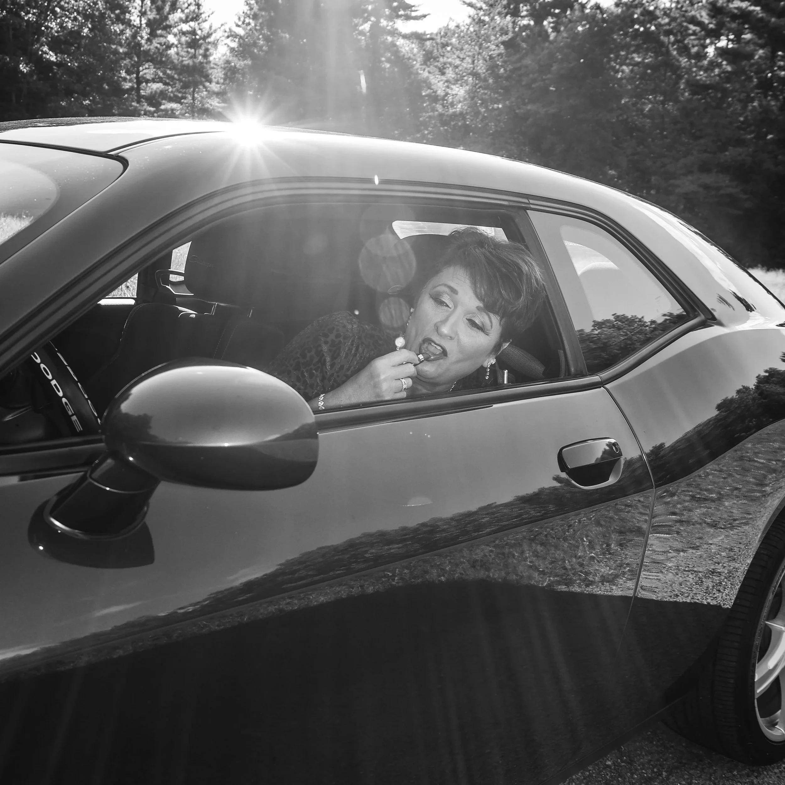 A woman with short hair and earrings is sitting in the driver's seat of a convertible car, applying lipstick while looking out of the window. Sunlight is shining from behind, creating lens flare, and trees are visible in the background.