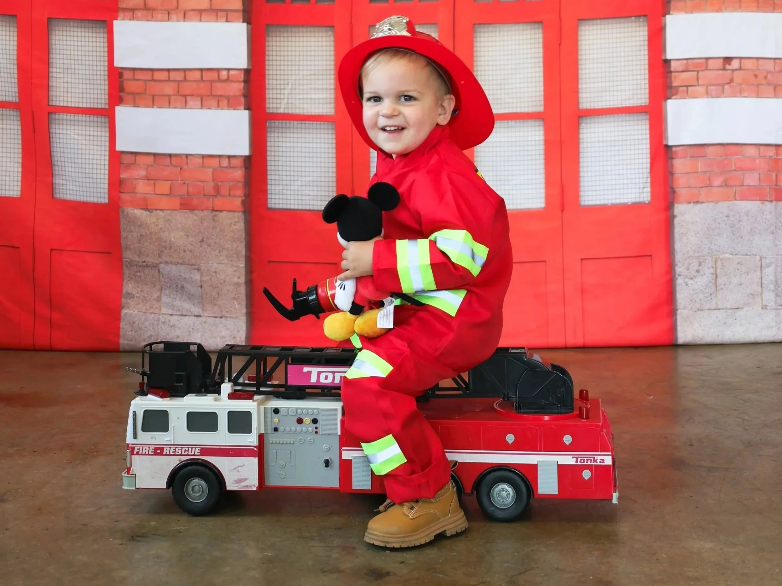 Child dressed as a firefighter sitting on a toy fire truck, holding a Mickey Mouse stuffed toy, with a red fire station backdrop in the background.