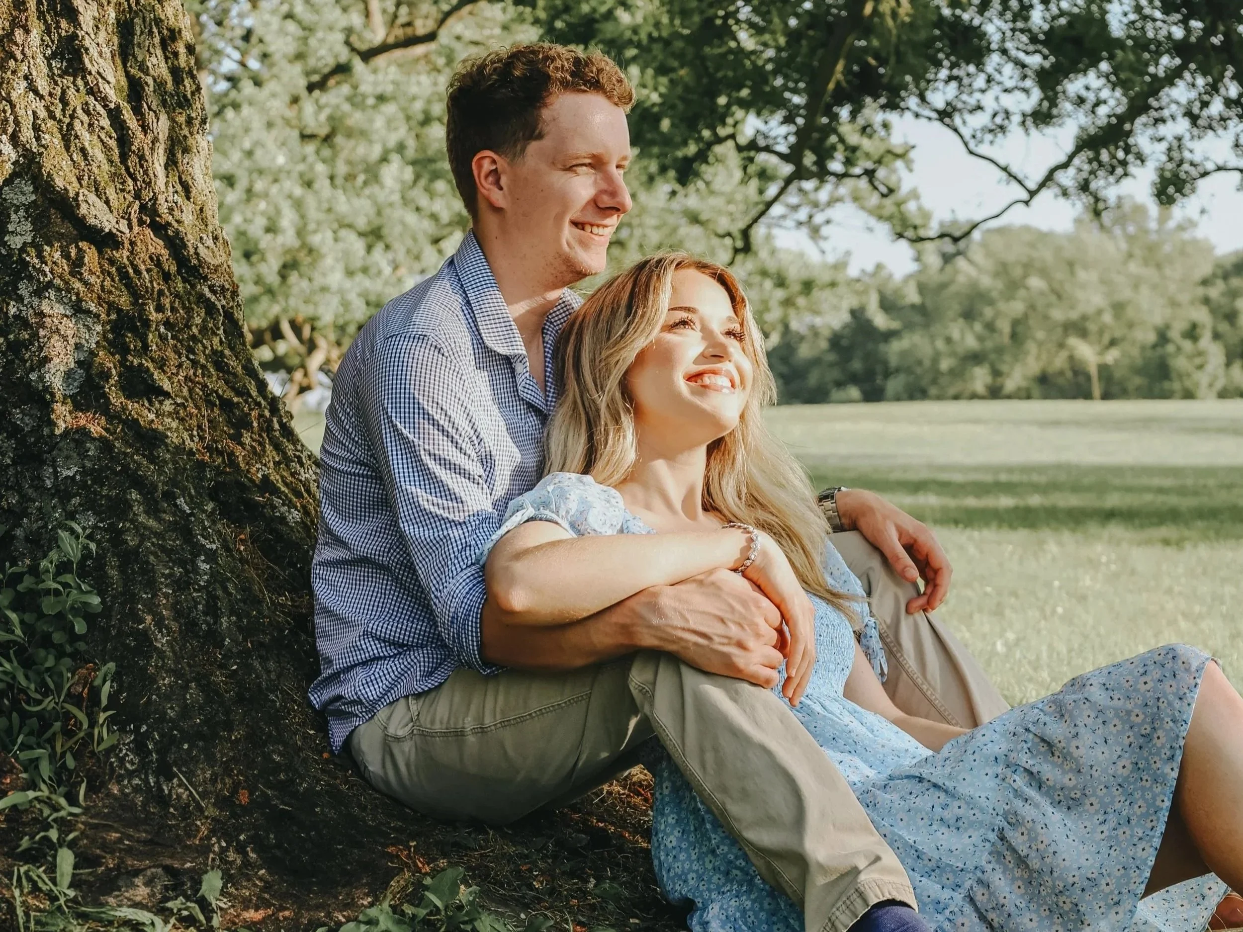 A smiling couple sitting against a tree outdoors on a sunny day, enjoying nature.