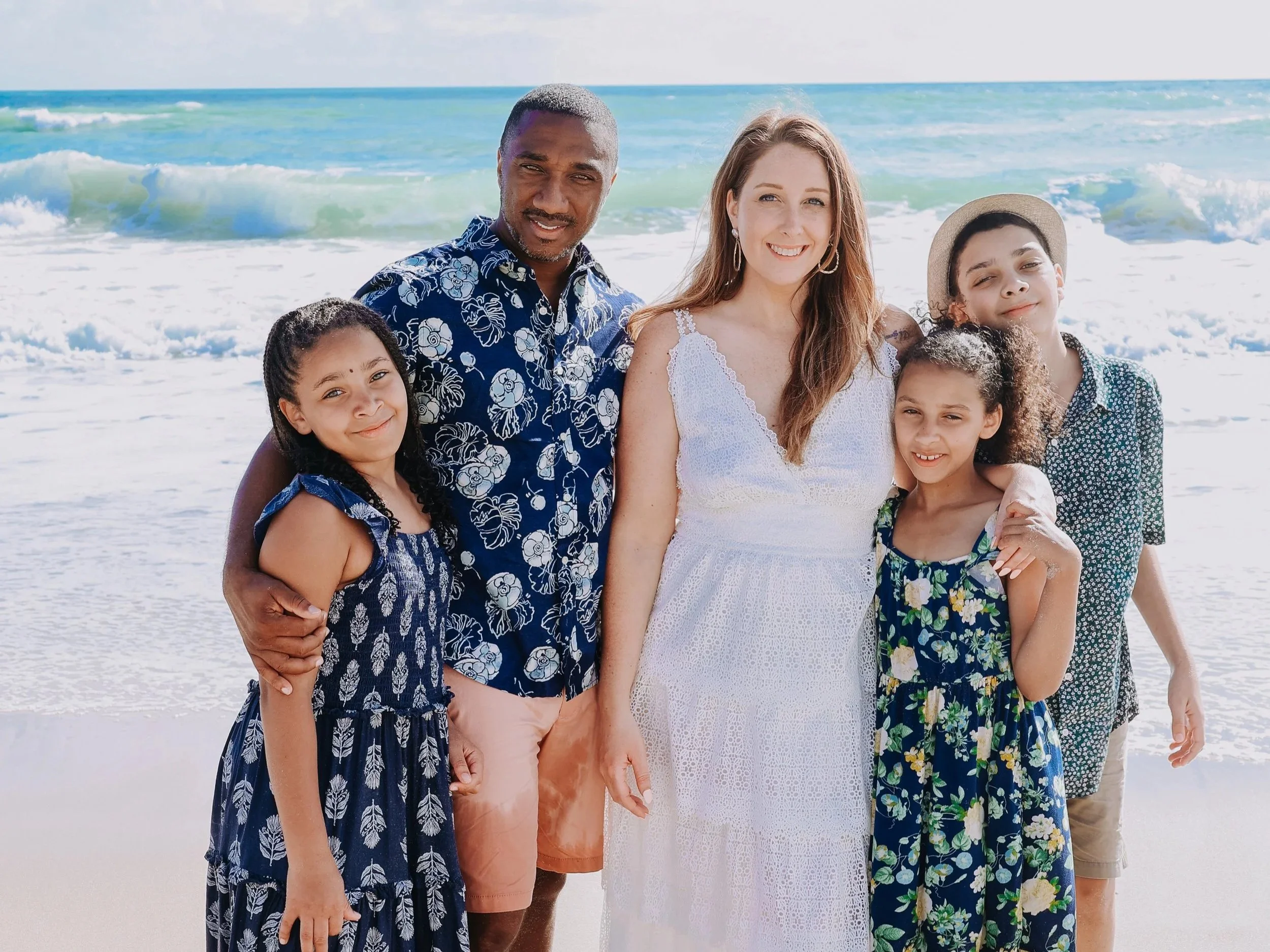 A family of six standing on a beach near the ocean, smiling at the camera. The group includes two adults and four children, dressed in summer clothing.