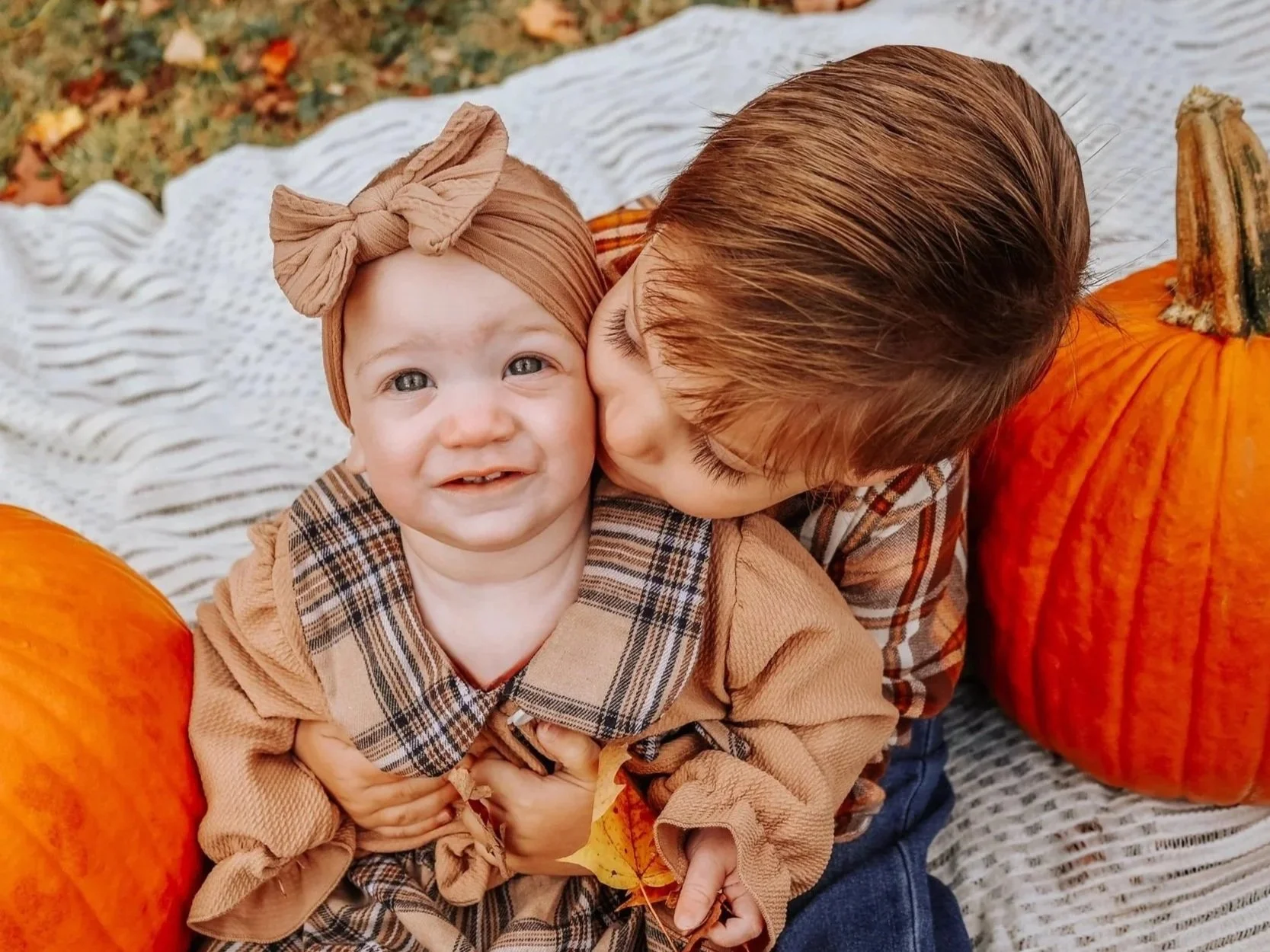 A young child with a headband kissing an older child on the cheek, seated between two large pumpkins outdoors on a blanket.