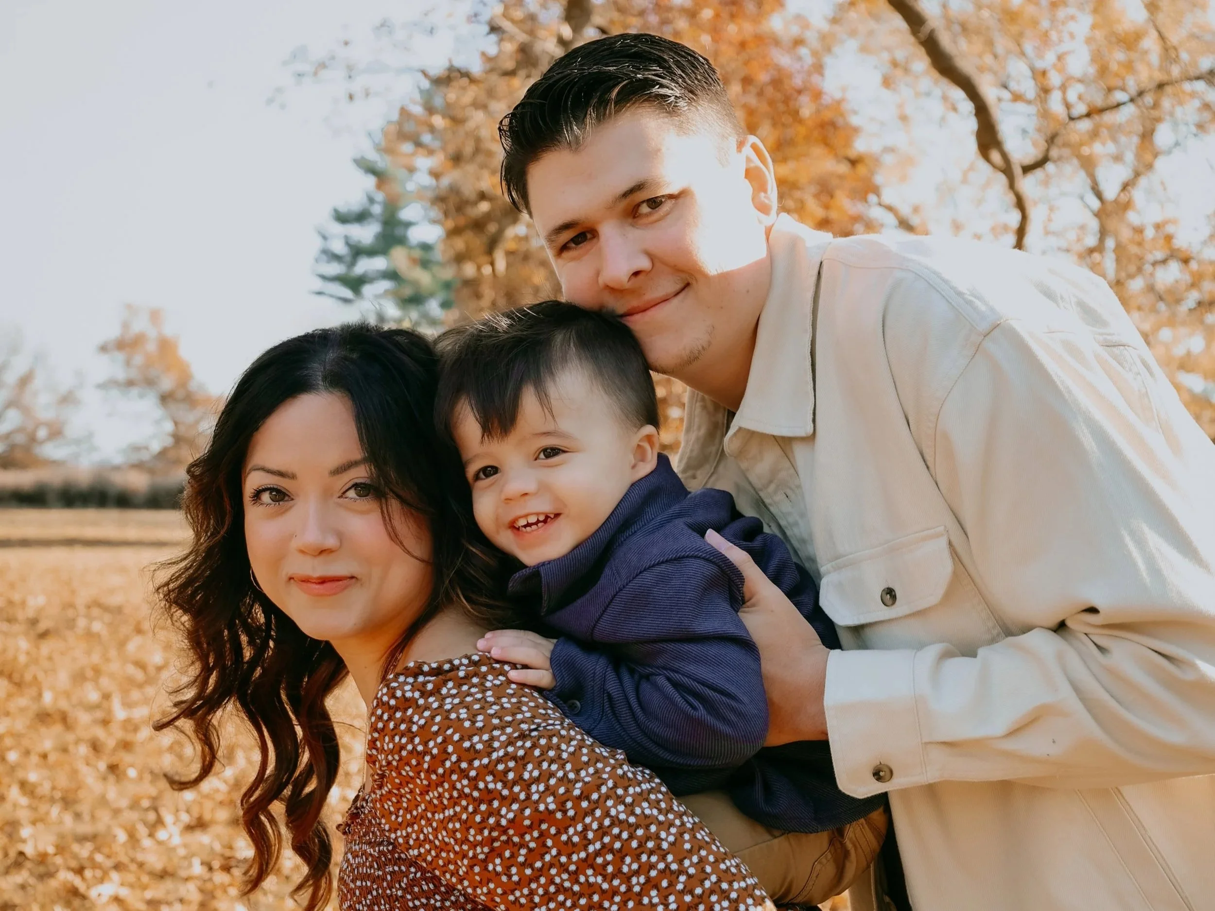 A family of three smiling outdoors during autumn, with golden leaves and trees in the background. The mother has dark wavy hair, the father has short dark hair, and the young boy has dark straight hair and is wearing a navy jacket.