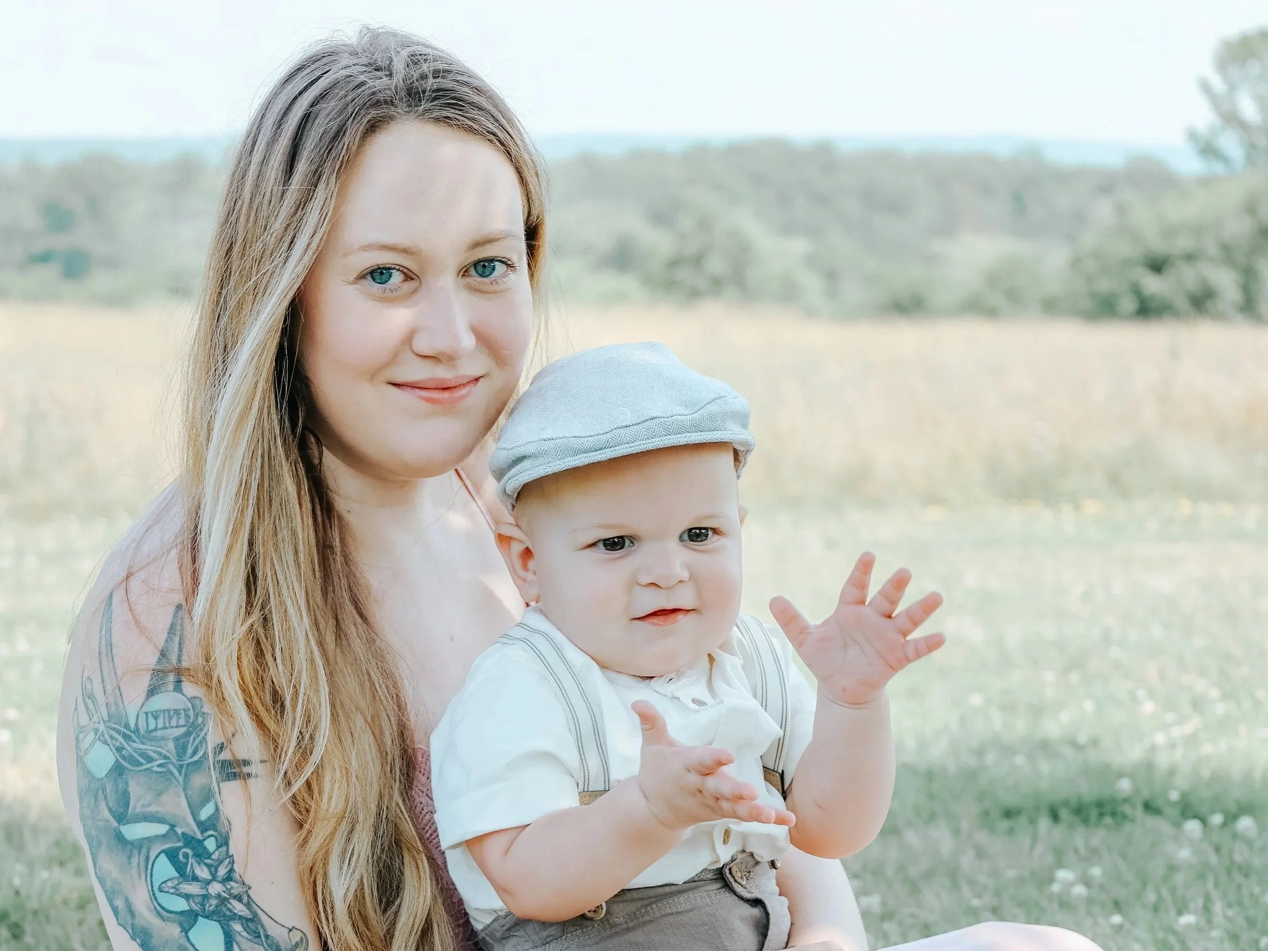 A woman with long blonde hair and a tattoo on her arm is holding a young child wearing a gray hat in a field with trees in the background.