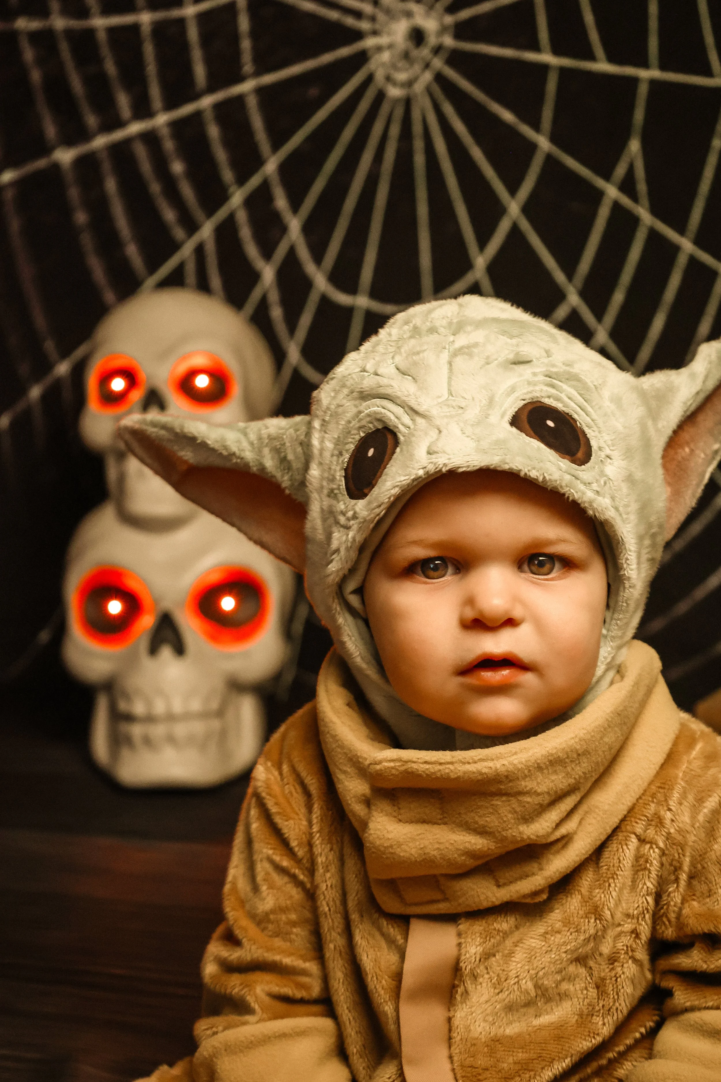 A young child dressed in a Yoda costume, with a hood featuring Yoda's face and ears, sitting indoors with Halloween-themed decorations behind them, including two glowing skulls with red LED eyes and a large spider web in the background.