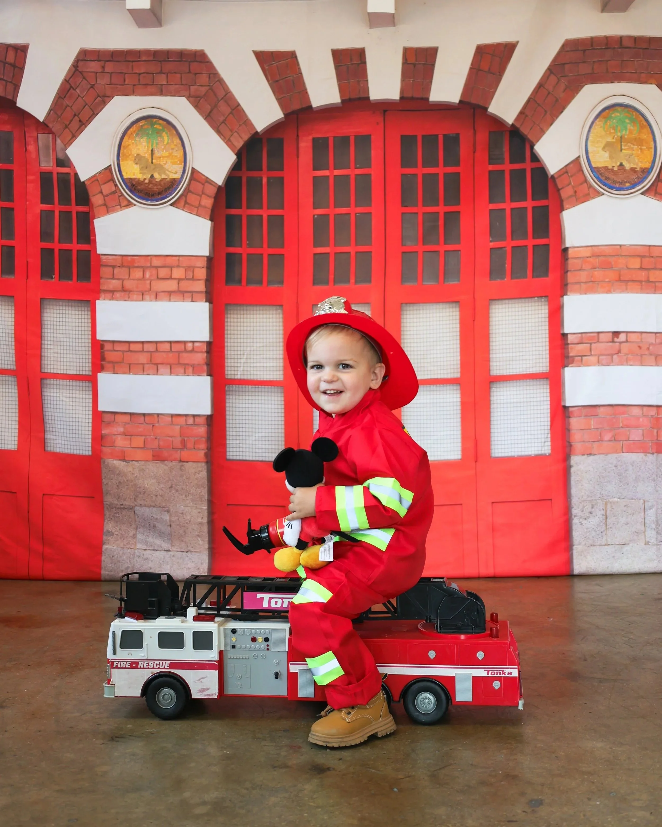 A young child dressed as a firefighter, sitting on a toy fire truck, holding a Mickey Mouse plush toy, with a red fire station backdrop.