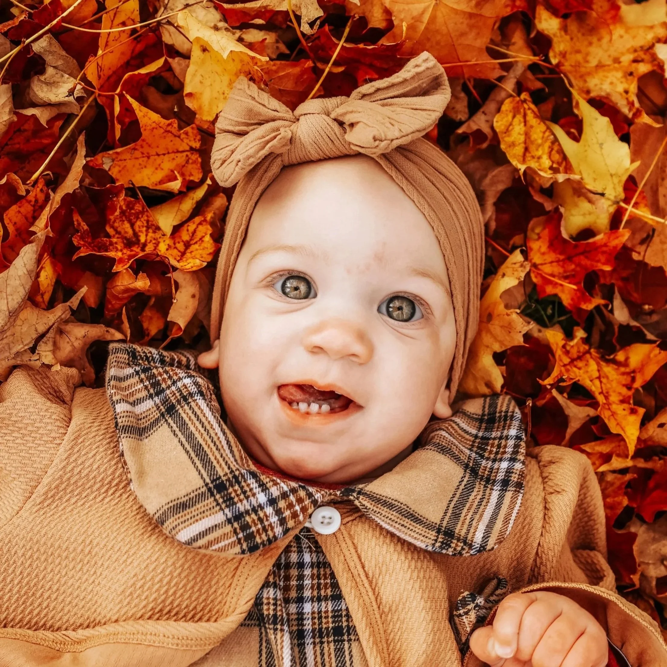 A smiling baby with blue eyes, lying on a bed of colorful autumn leaves, wearing a tan headwrap with a bow and a tan plaid coat.