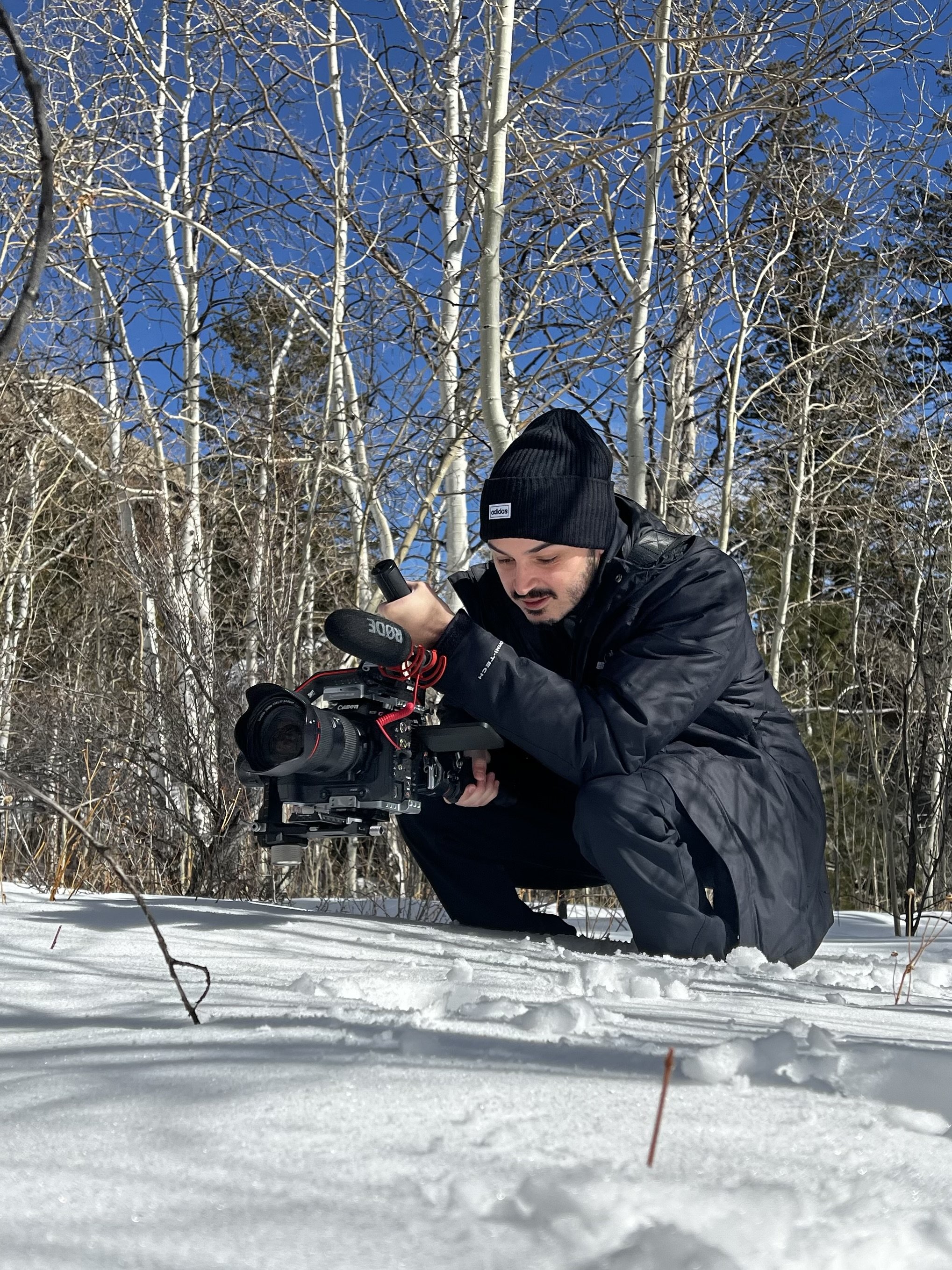 Person crouching in snowy forest holding a professional camera, framed by leafless trees and a clear blue sky.