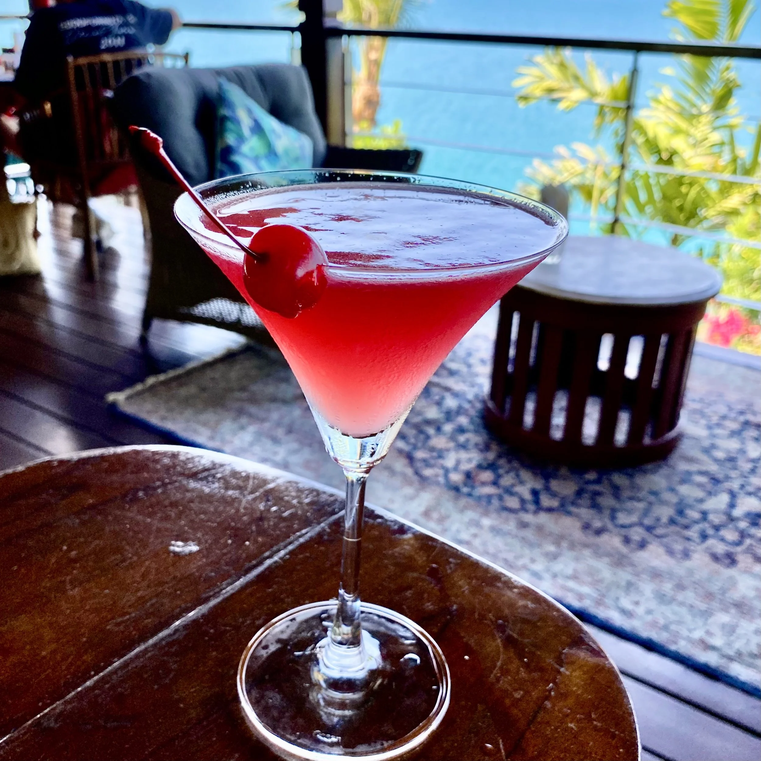 A pink cocktail with a cherry and an umbrella on a wooden table near a balcony overlooking water, with outdoor furniture and greenery in the background.