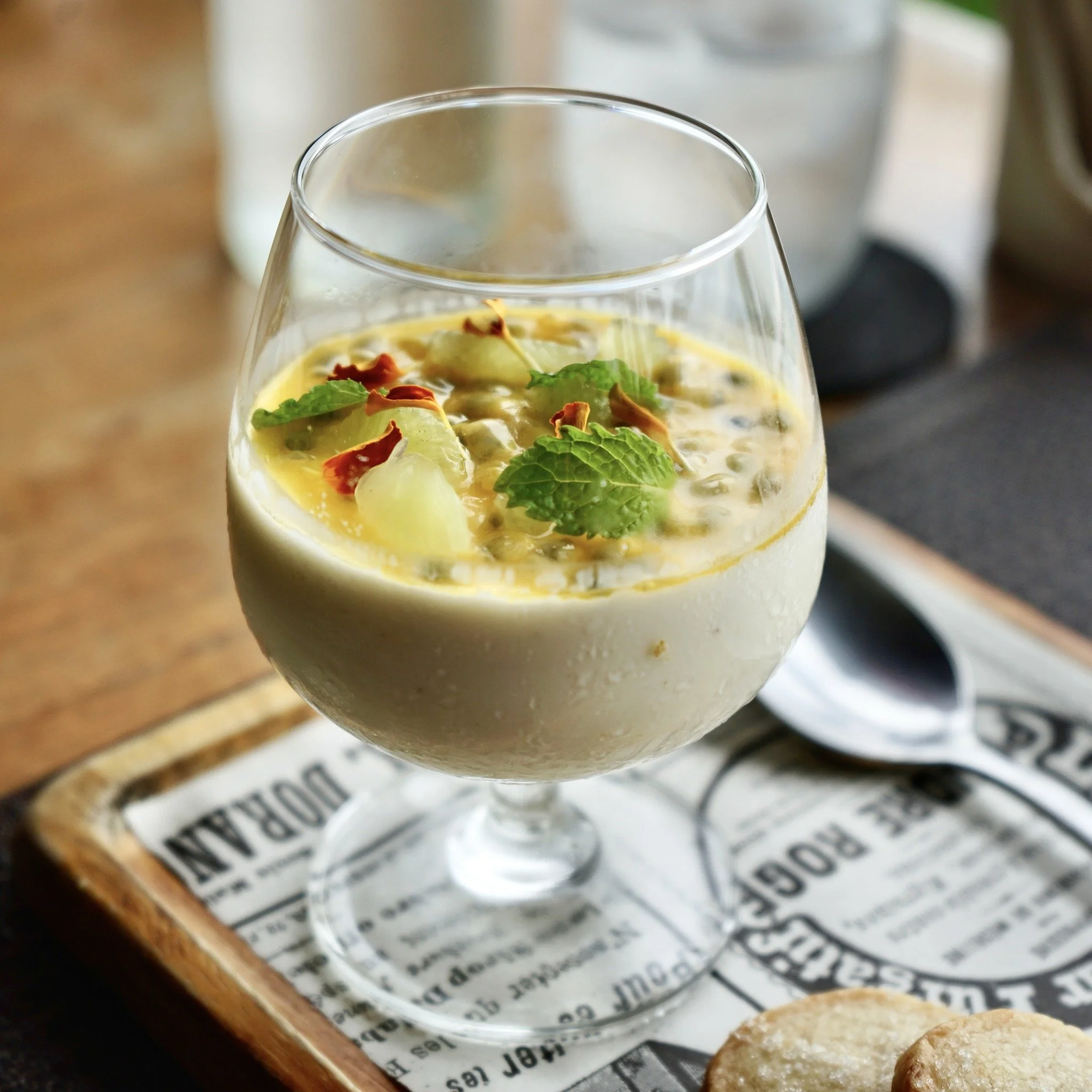 Close-up of a glass with a creamy yellow dessert topped with fresh mint leaves, sliced fruit, and dried flower petals, placed on a sheet of newspaper on a wooden surface.