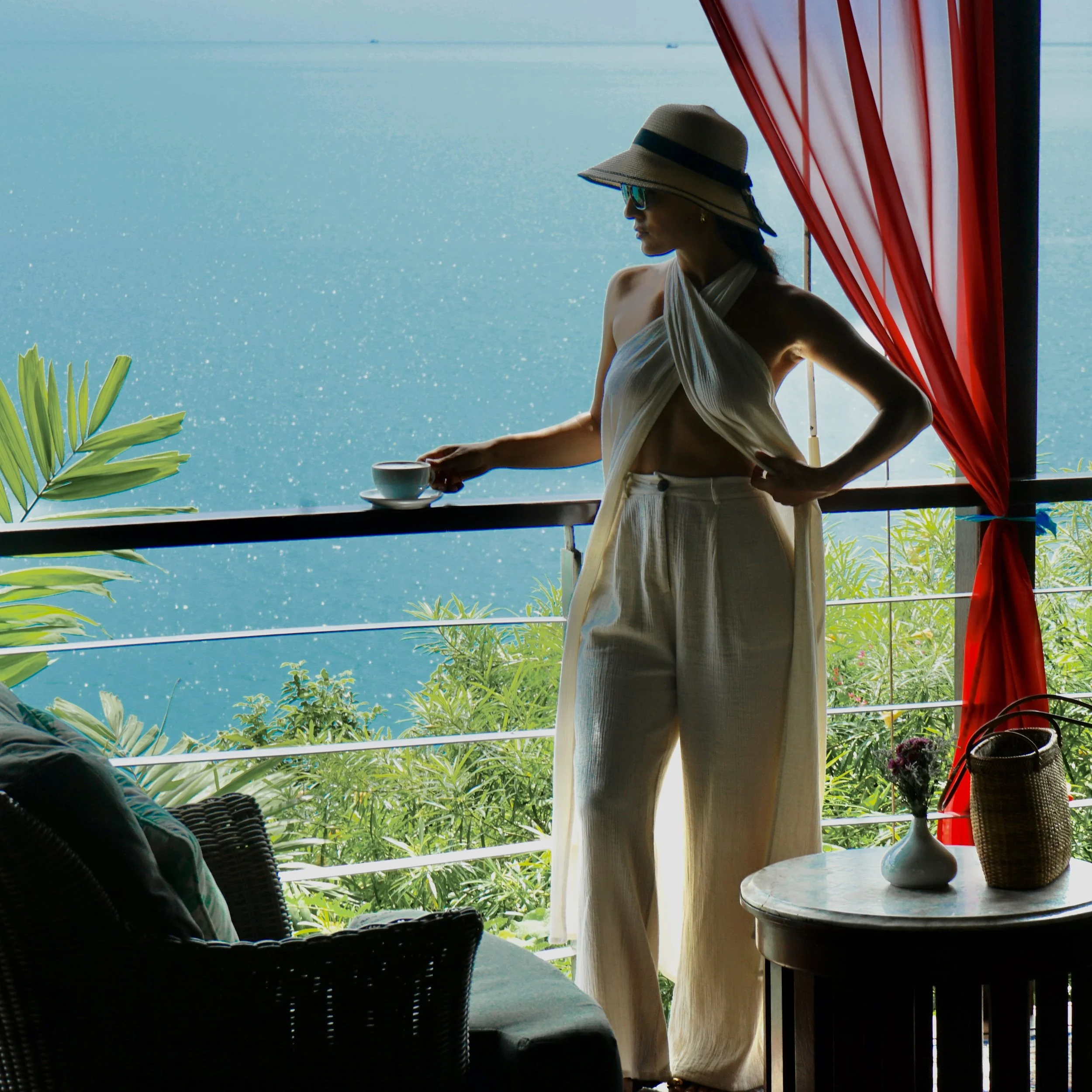 A woman is standing on a balcony overlooking the ocean, wearing a wide-brimmed hat and sunglasses, with a drink in her hand. The scene includes tropical plants, a table with decorative items, and bright red curtains.