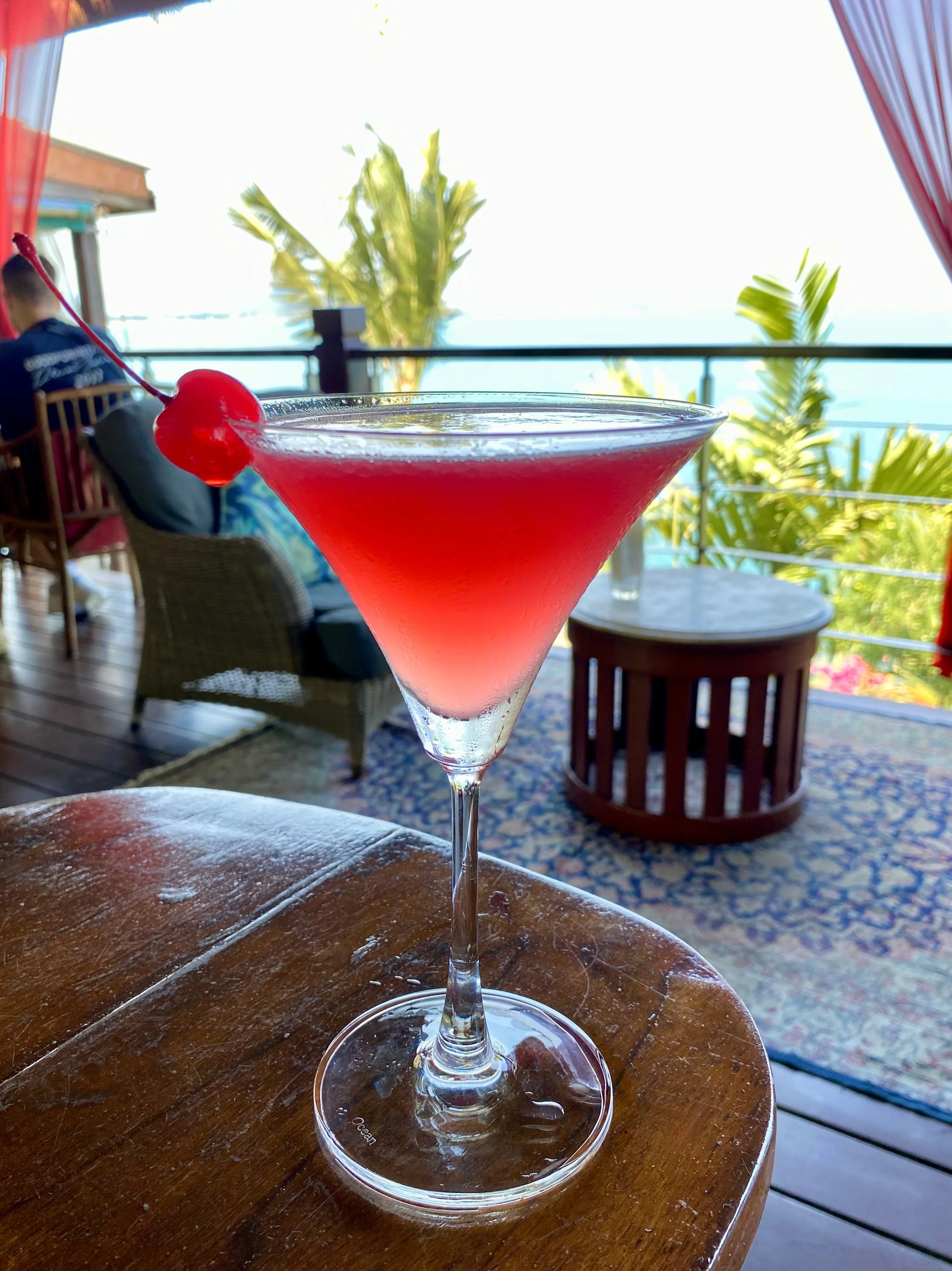 A pink cocktail with a cherry garnish on a wooden table at a seaside outdoor restaurant, with palm trees, a railing, colorful curtains, a person sitting in the background, and a view of the ocean.