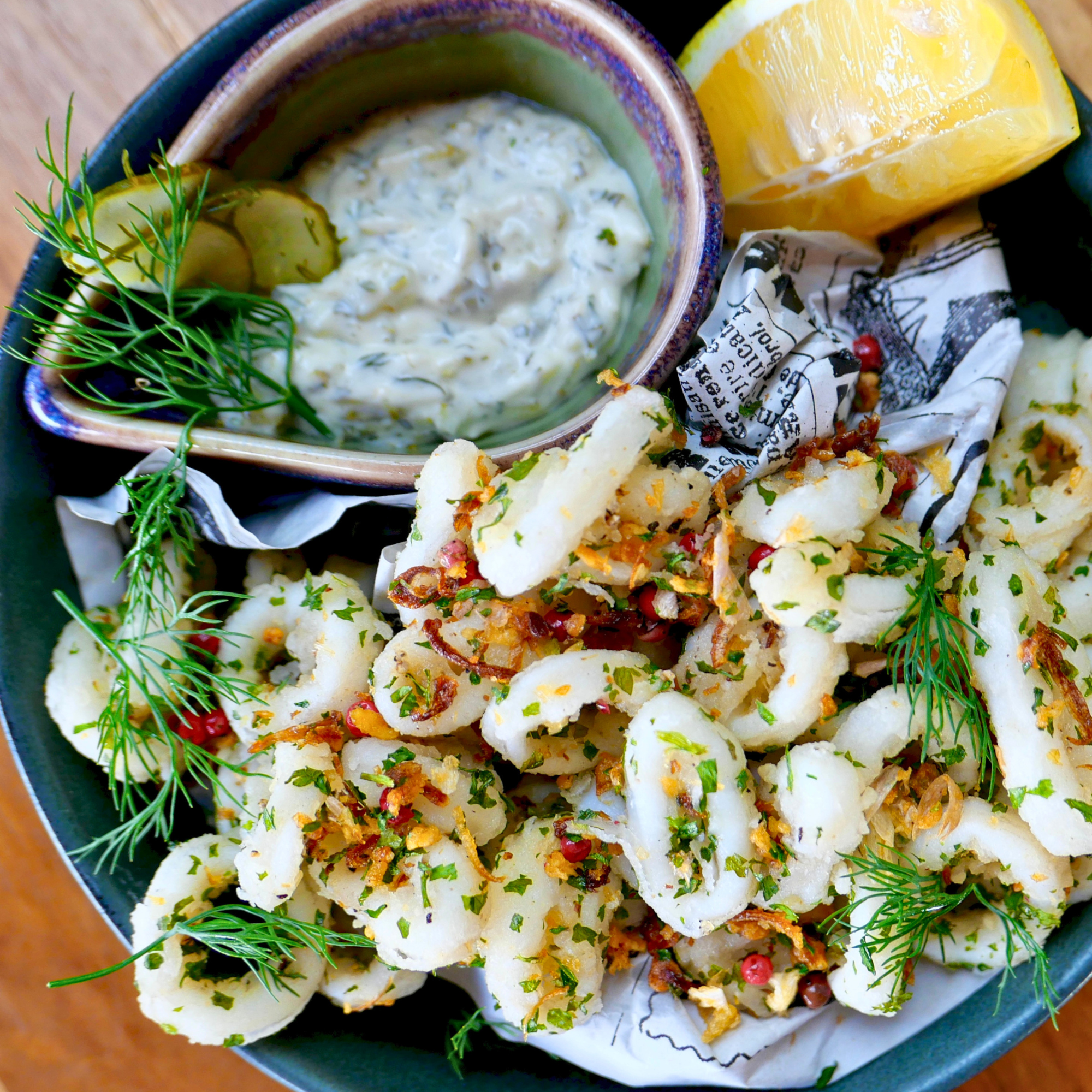 Close-up of a plate with sliced cauliflower topped with herbs and spices, garnished with fresh dill, accompanied by lemon wedges, a small bowl of creamy dip, and crumpled newspaper-style paper.
