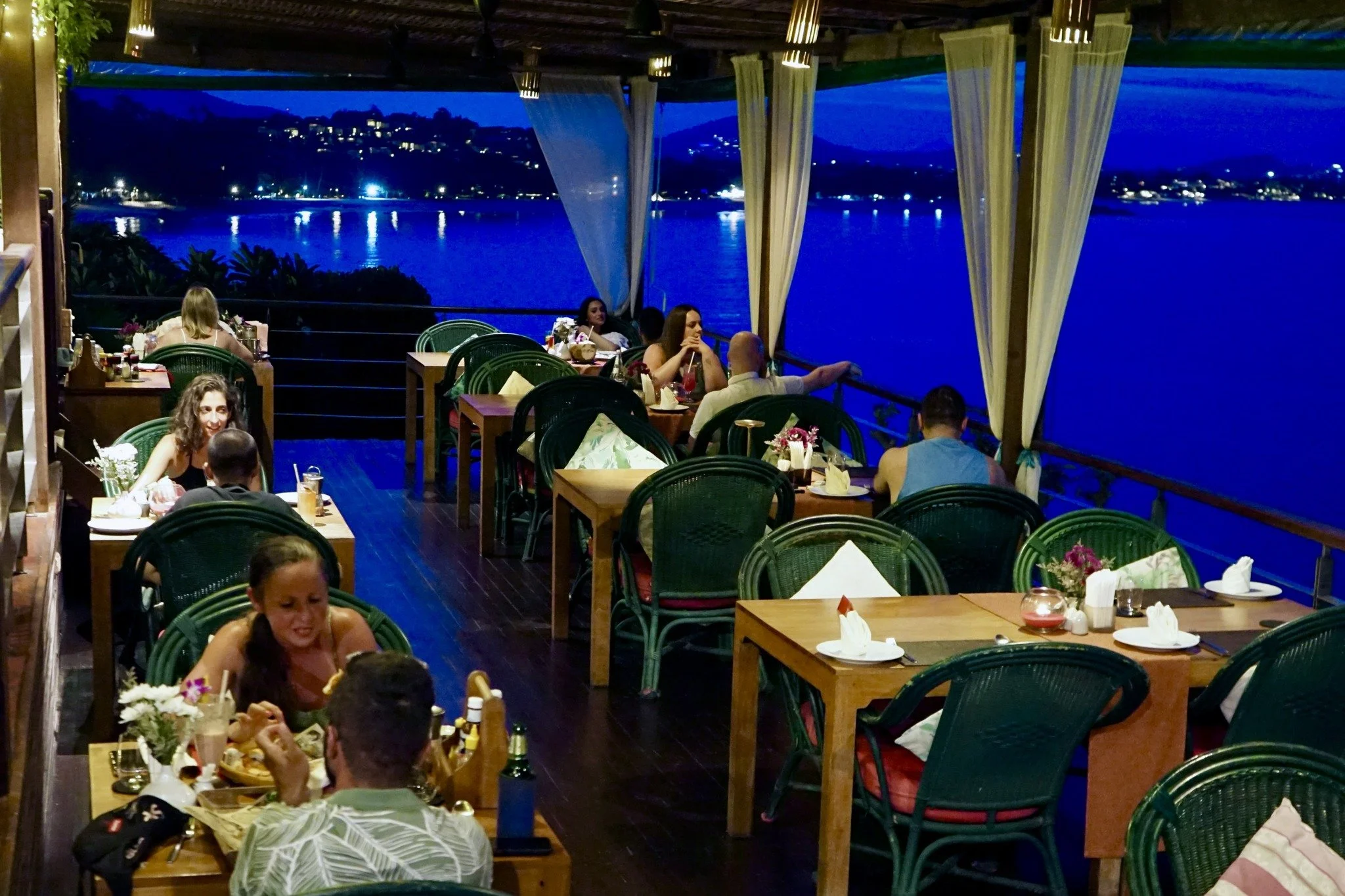 A nighttime scene of a restaurant with outdoor seating by the water. Several tables with people dining, overlooking a body of water with lights reflecting on it, and a hillside with houses visible in the background. The restaurant has wooden flooring