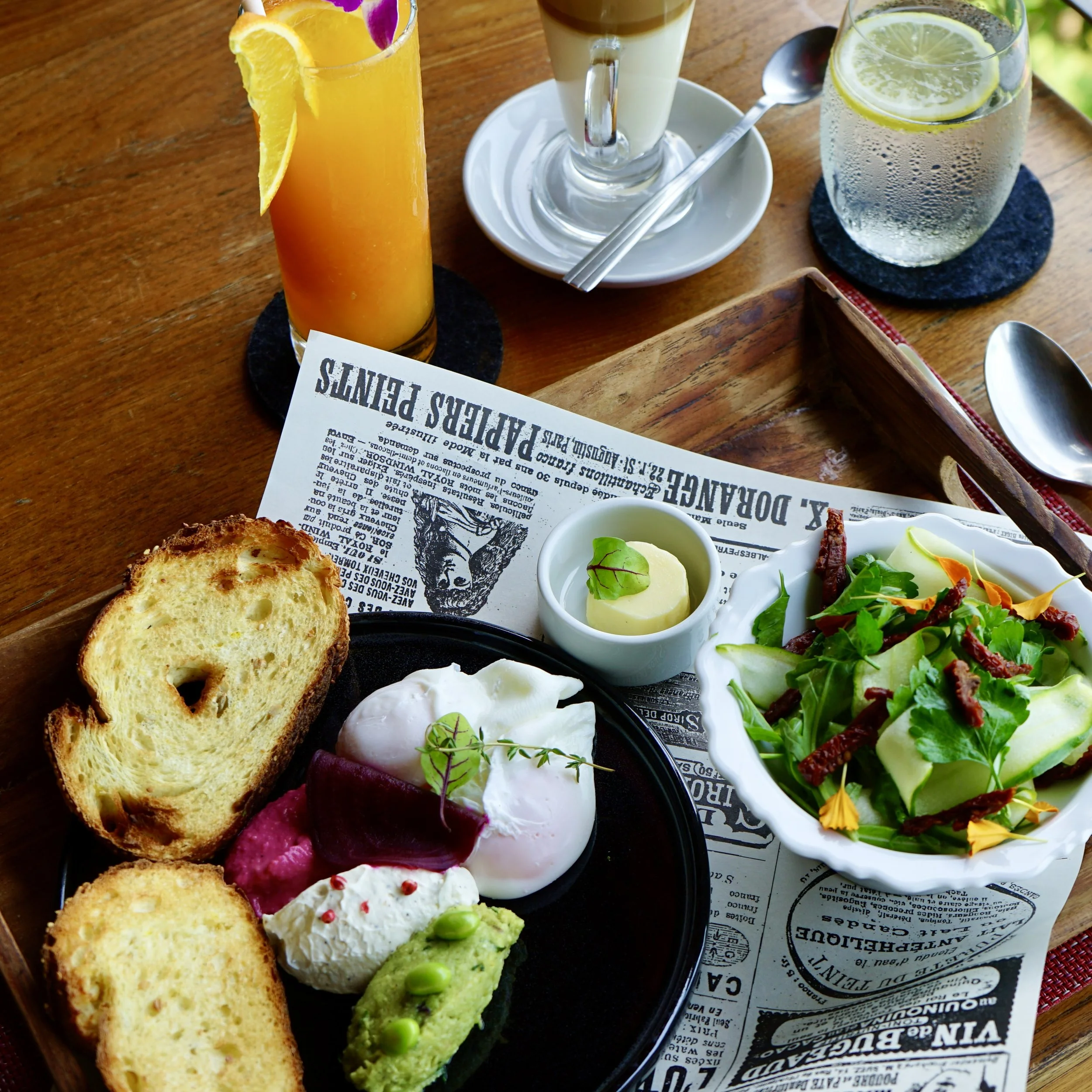 Three beverages: a tall glass of orange juice with orange slices and an orchid flower, a layered coffee drink in a glass mug, and a glass of sparkling water with lemon slices, all on a wooden table. There is also a plate with assorted food including 