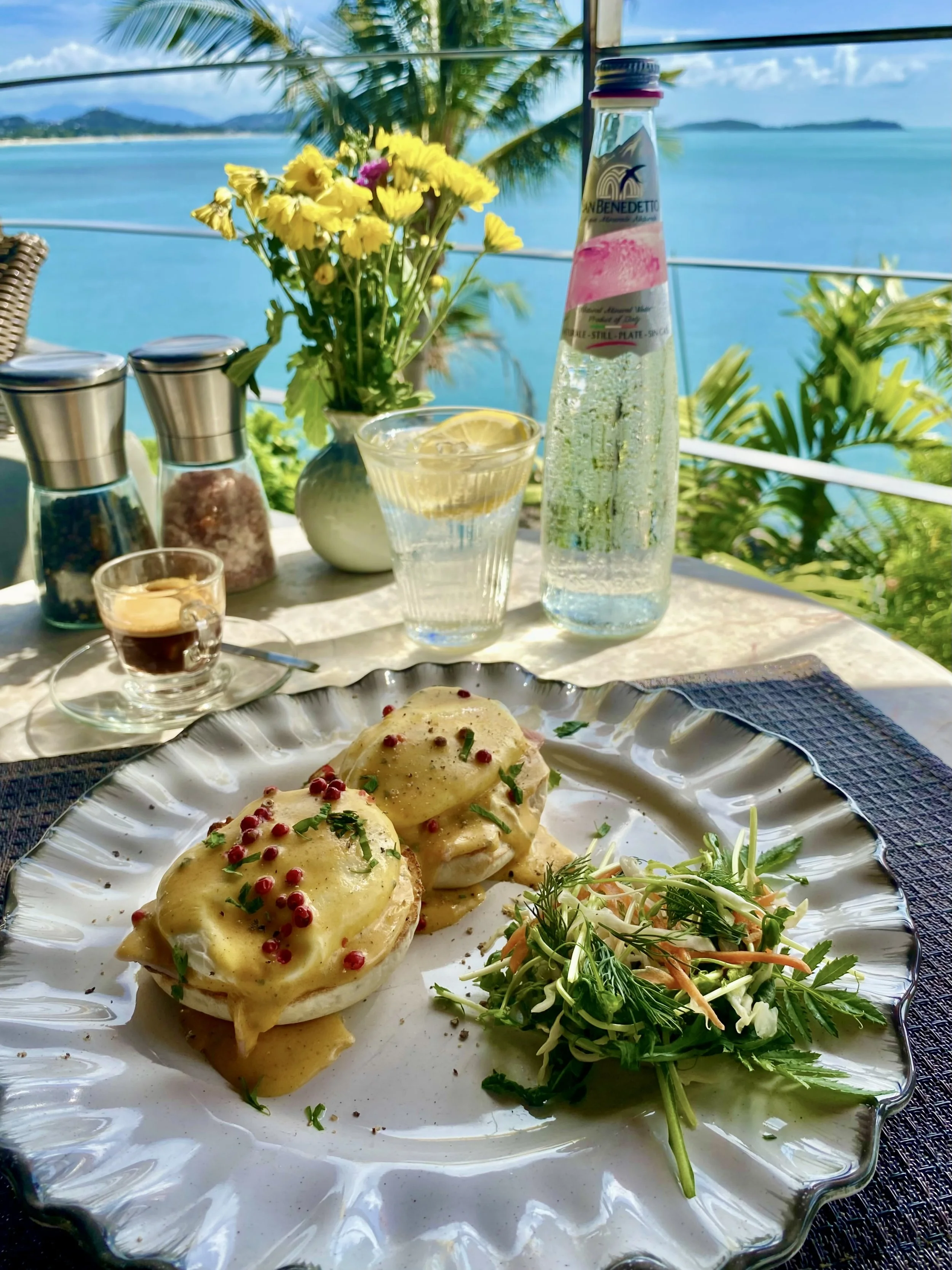 A plate of eggs benedict topped with hollandaise sauce, garnished with pink peppercorns and chives, served with a side salad with herbs and vegetables, on a table in a seaside restaurant with a view of the ocean and tropical plants.