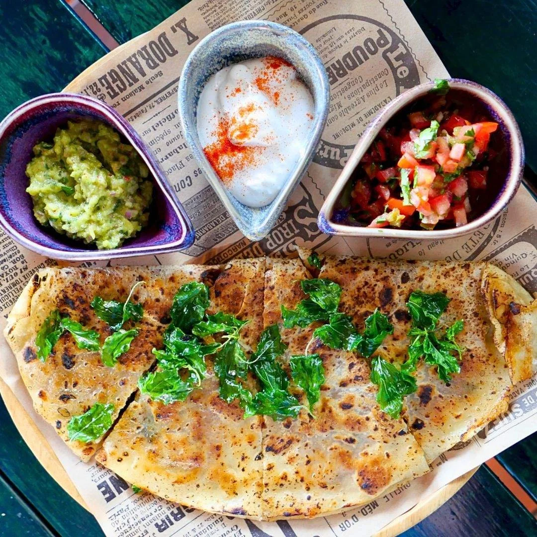 A plate of flatbread garnished with green herbs, accompanied by three small bowls of guacamole, sour cream with paprika, and pico de gallo salsa, on top of a newspaper-lined wooden table.