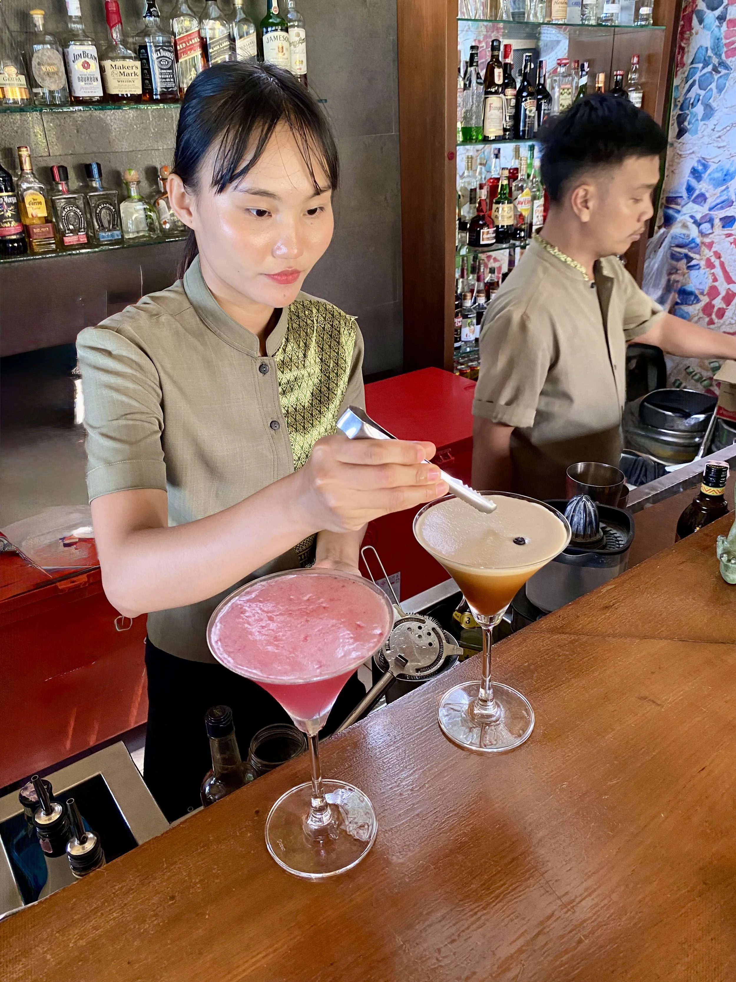 A woman making cocktails at a bar with two colorful drinks in martini glasses in the foreground and two bartenders in the background, along with liquor bottles on shelves.