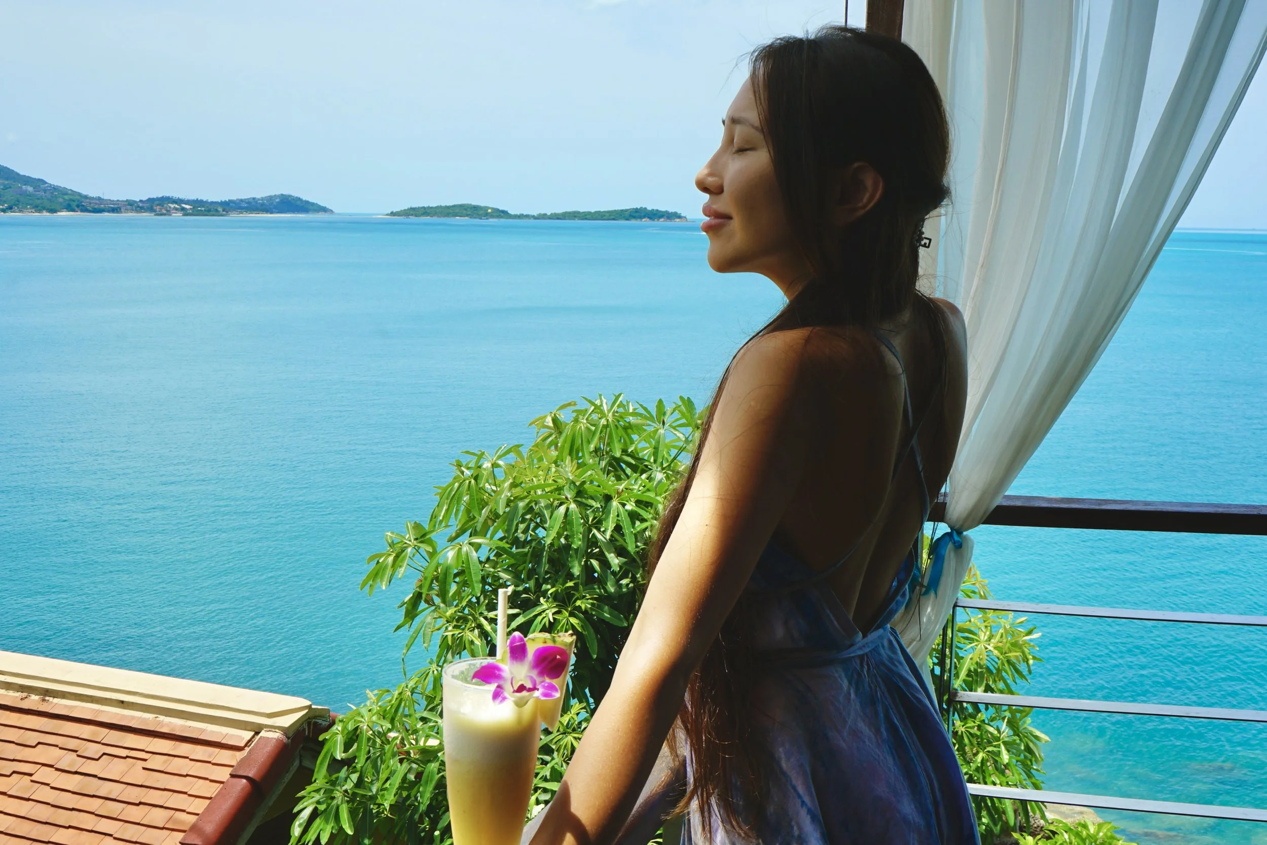 A woman with long dark hair and closed eyes standing on a balcony overlooking the ocean, with a drink garnished with a pink flower in the foreground.