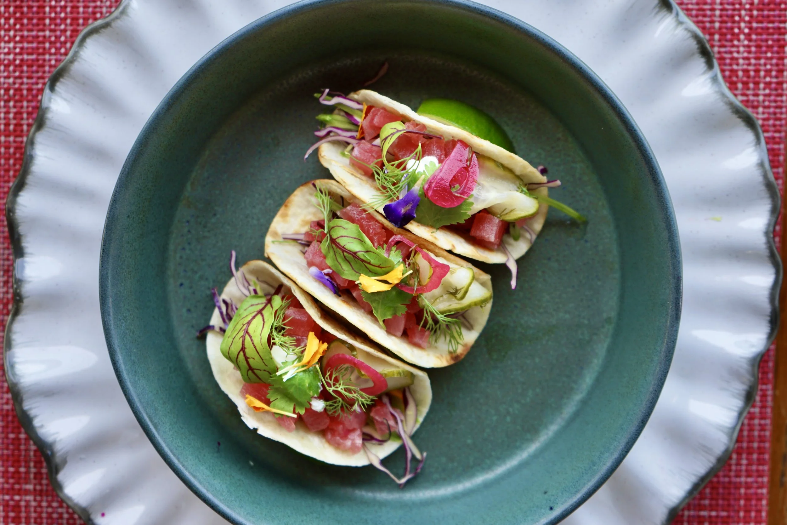 Tacos filled with diced meat, fresh herbs, cucumber slices, and microgreens, served on a teal plate with a scalloped white edge, placed on a red woven placemat.