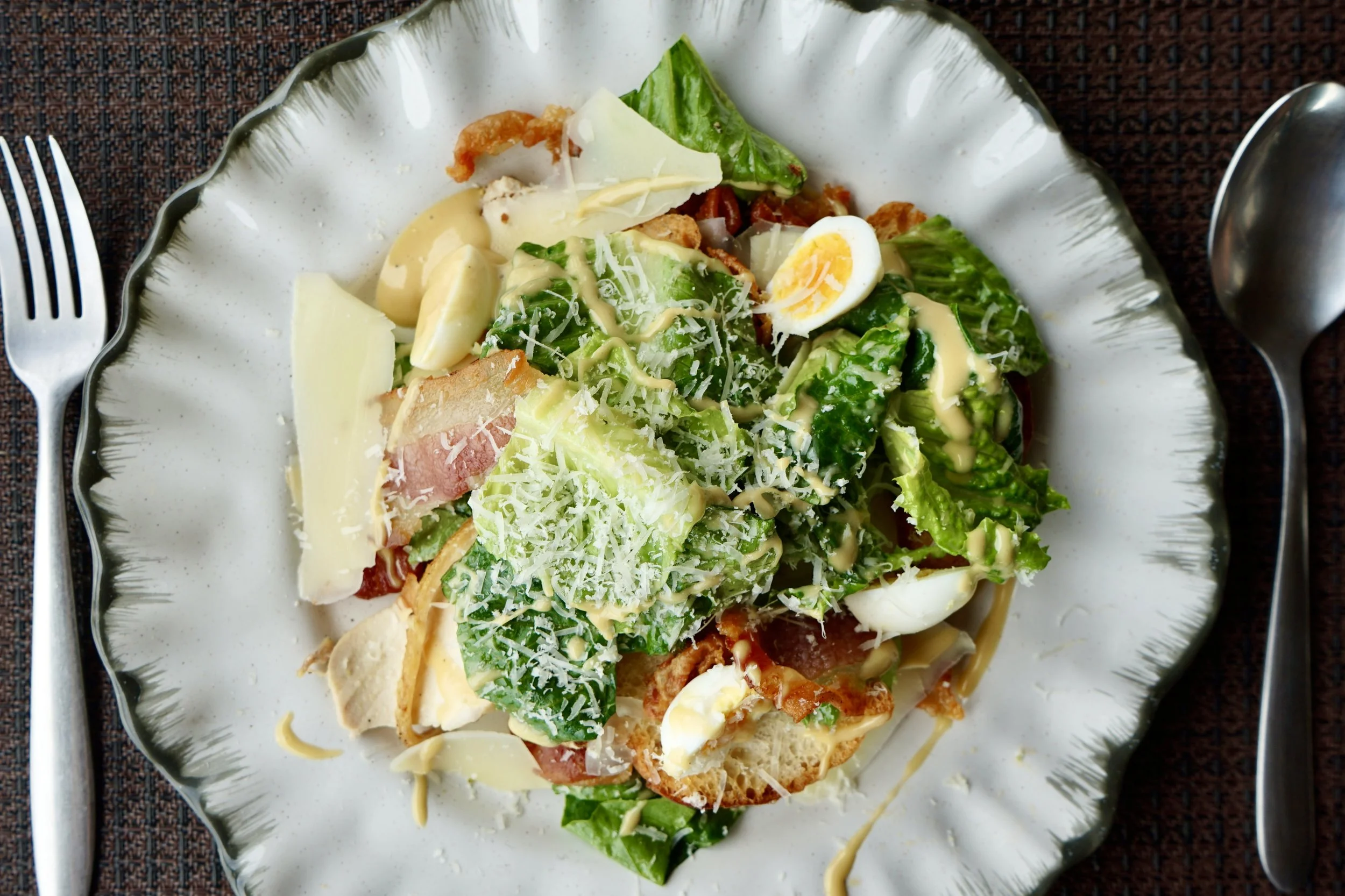 A plate of salad with lettuce, sliced boiled egg, shredded cheese, croutons, and creamy salad dressing, served on a decorative ceramic dish with fork and spoon beside it.