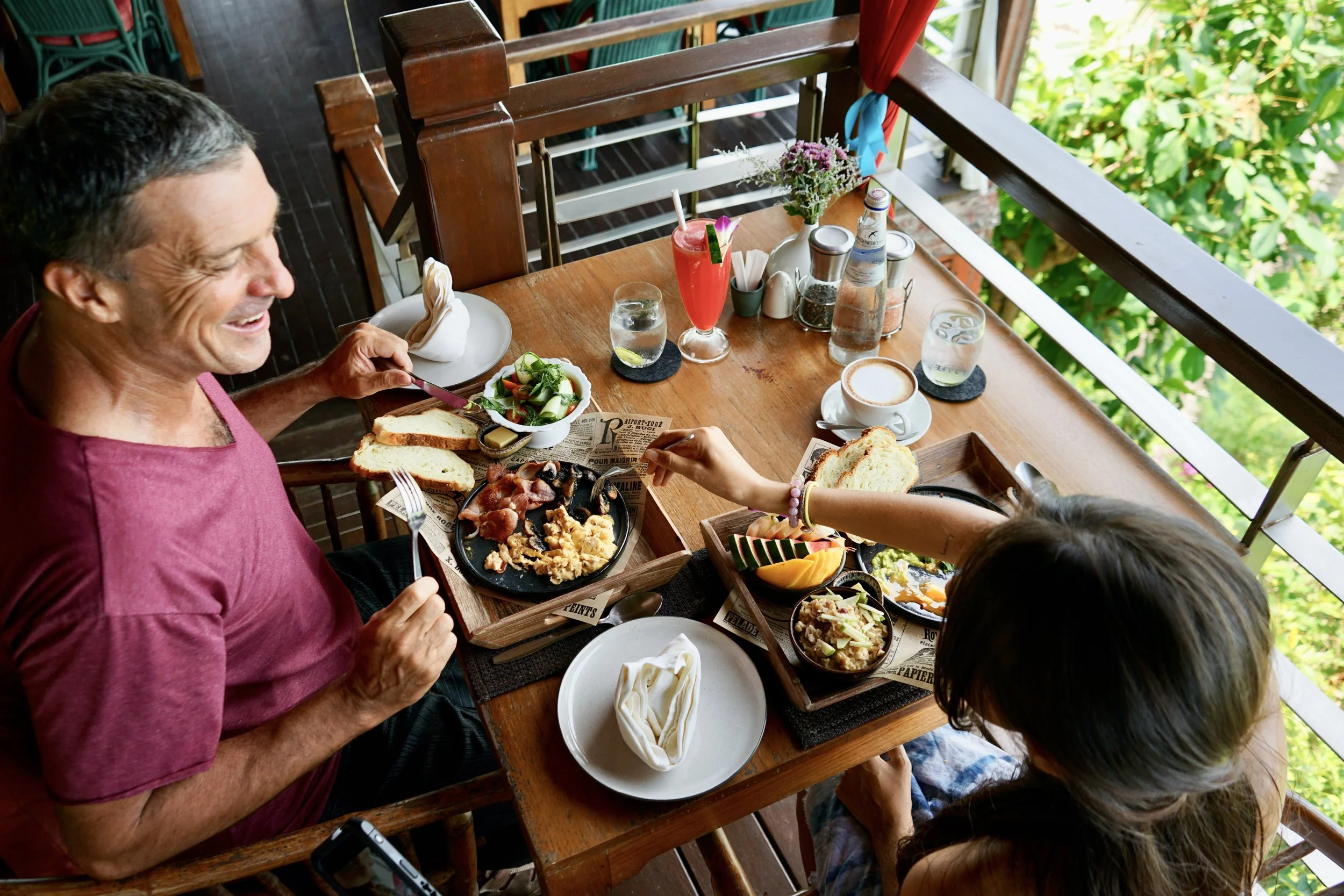 A man with gray hair smiling and eating at a wooden table with two children, surrounded by various dishes, drinks, and flowers on an outdoor balcony with green foliage visible outside.