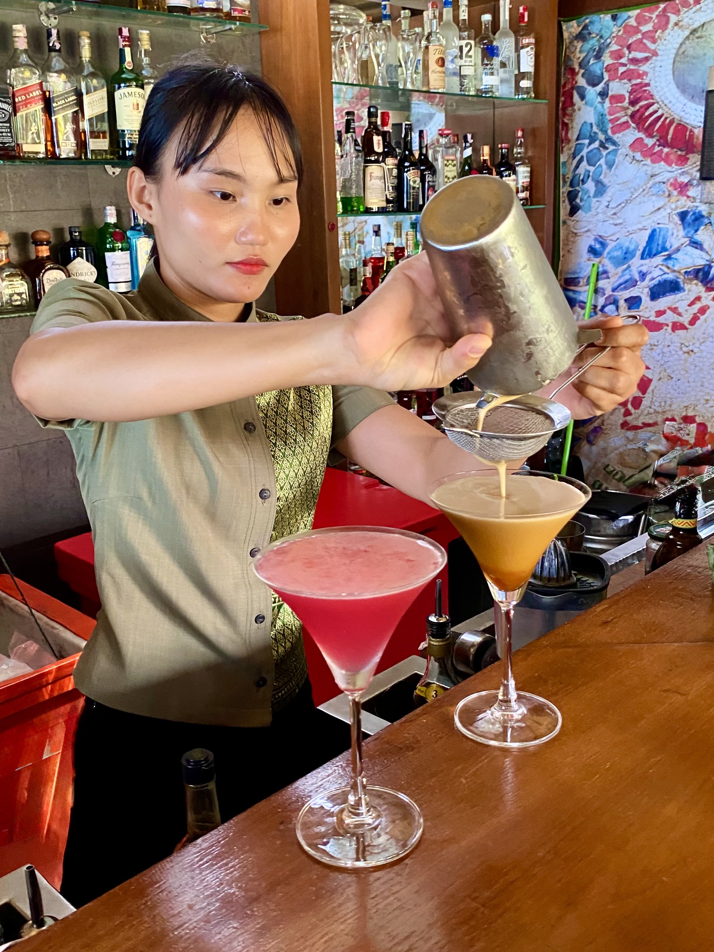 A woman pouring a cocktail through a fine mesh strainer into a martini glass, with two colorful cocktails on the bar counter in front of her.