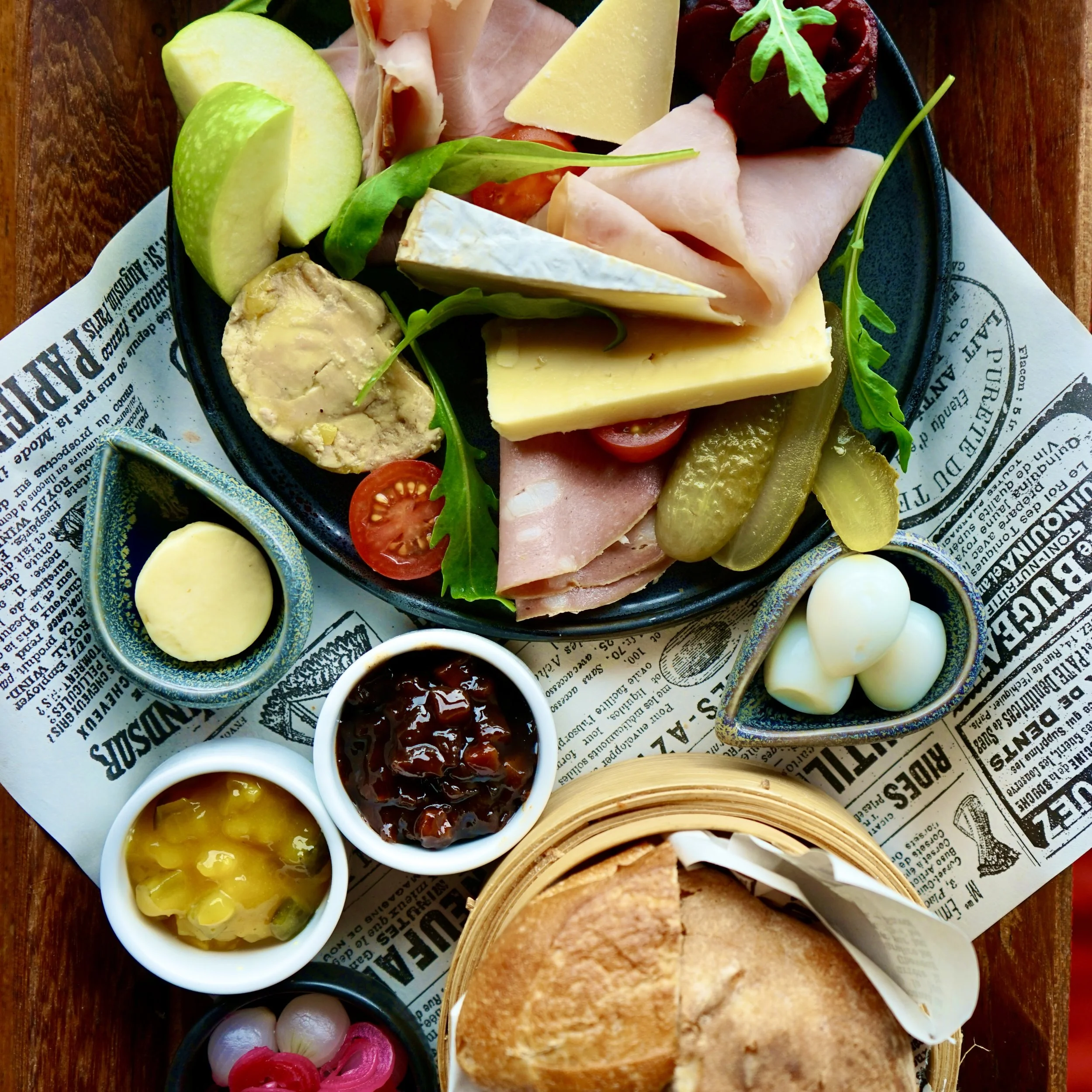 A black plate with cheeses, sliced meats, cherry tomatoes, pickles, green apple slices, arugula, and garnishes on a wooden table with newspaper-style print.