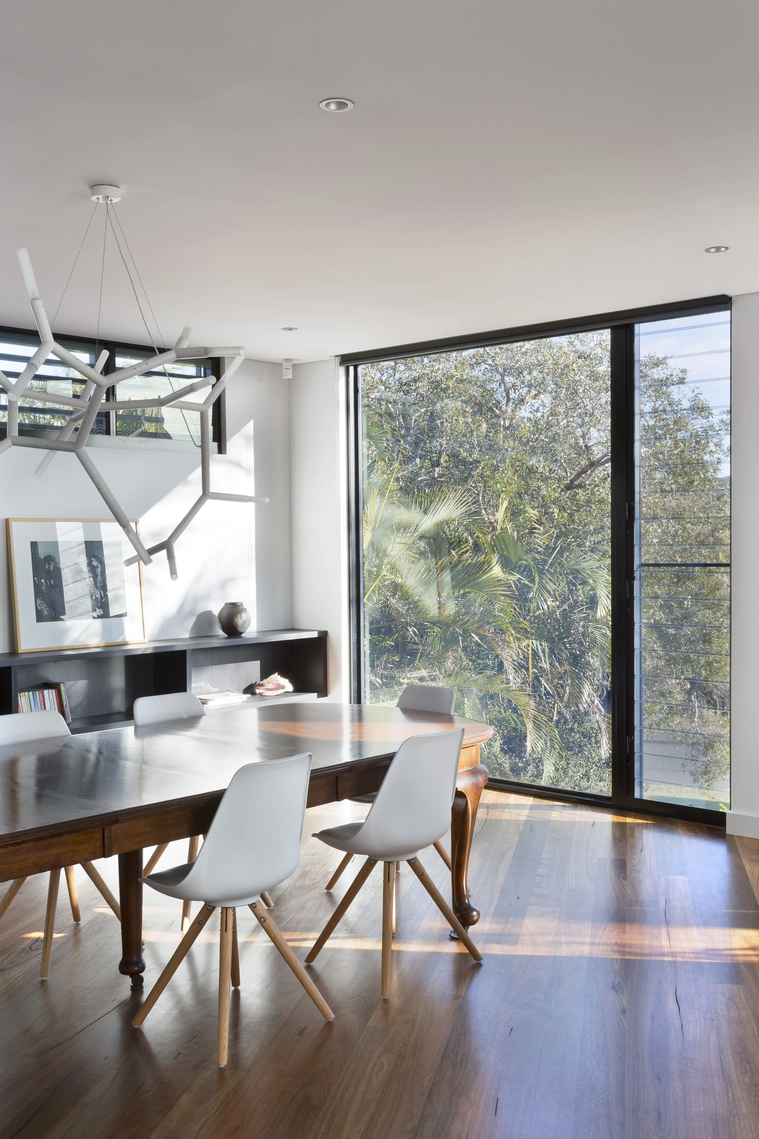 Modern dining room with wooden table and white chairs, large windows showing lush green trees outside, minimalist pendant light, and framed artwork on the wall.