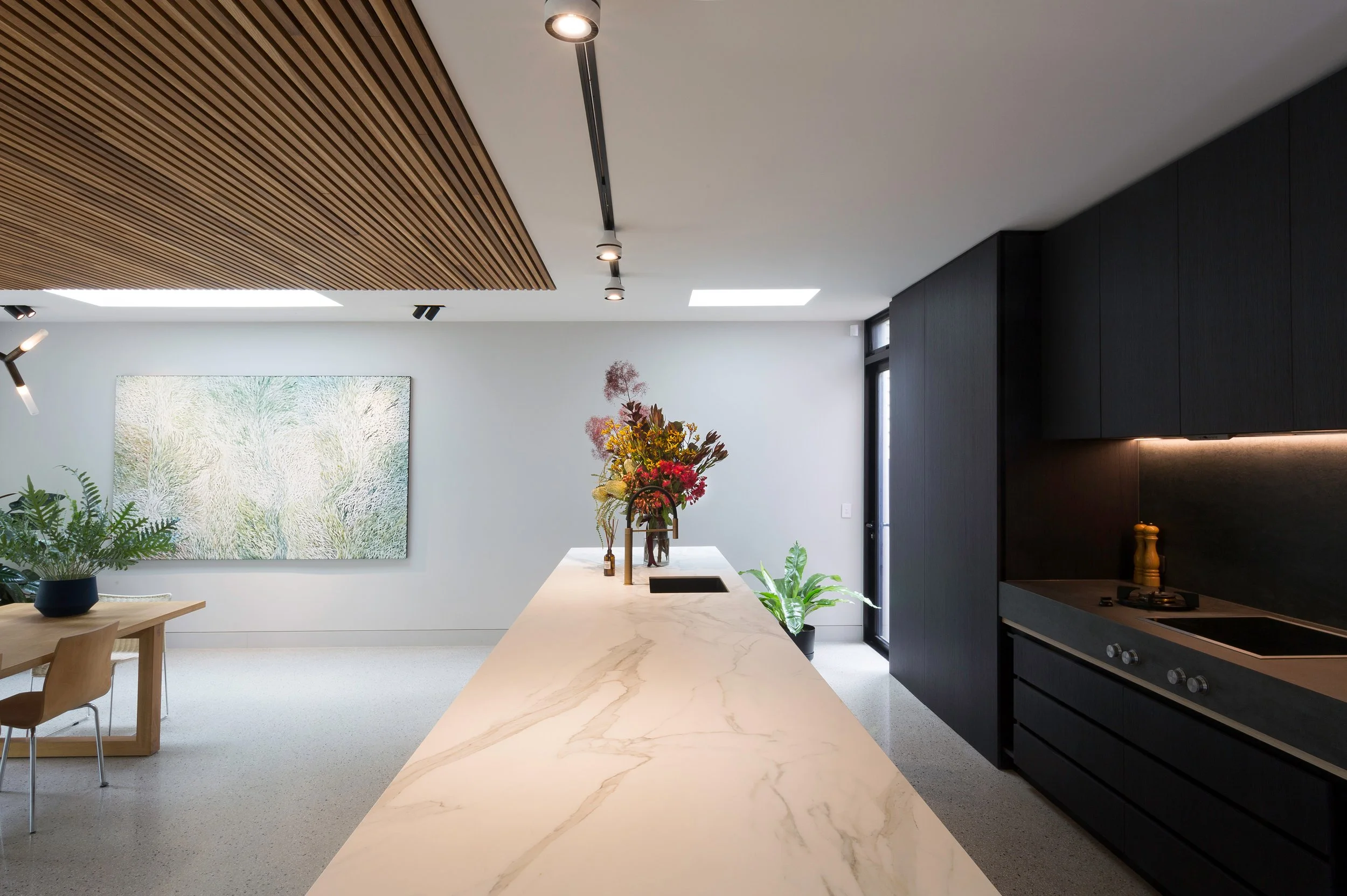 Modern kitchen with white marble island, black cabinetry, and wooden ceiling panels. There are plants and a colorful bouquet on the island, with a large abstract painting on the wall and a window letting in natural light.