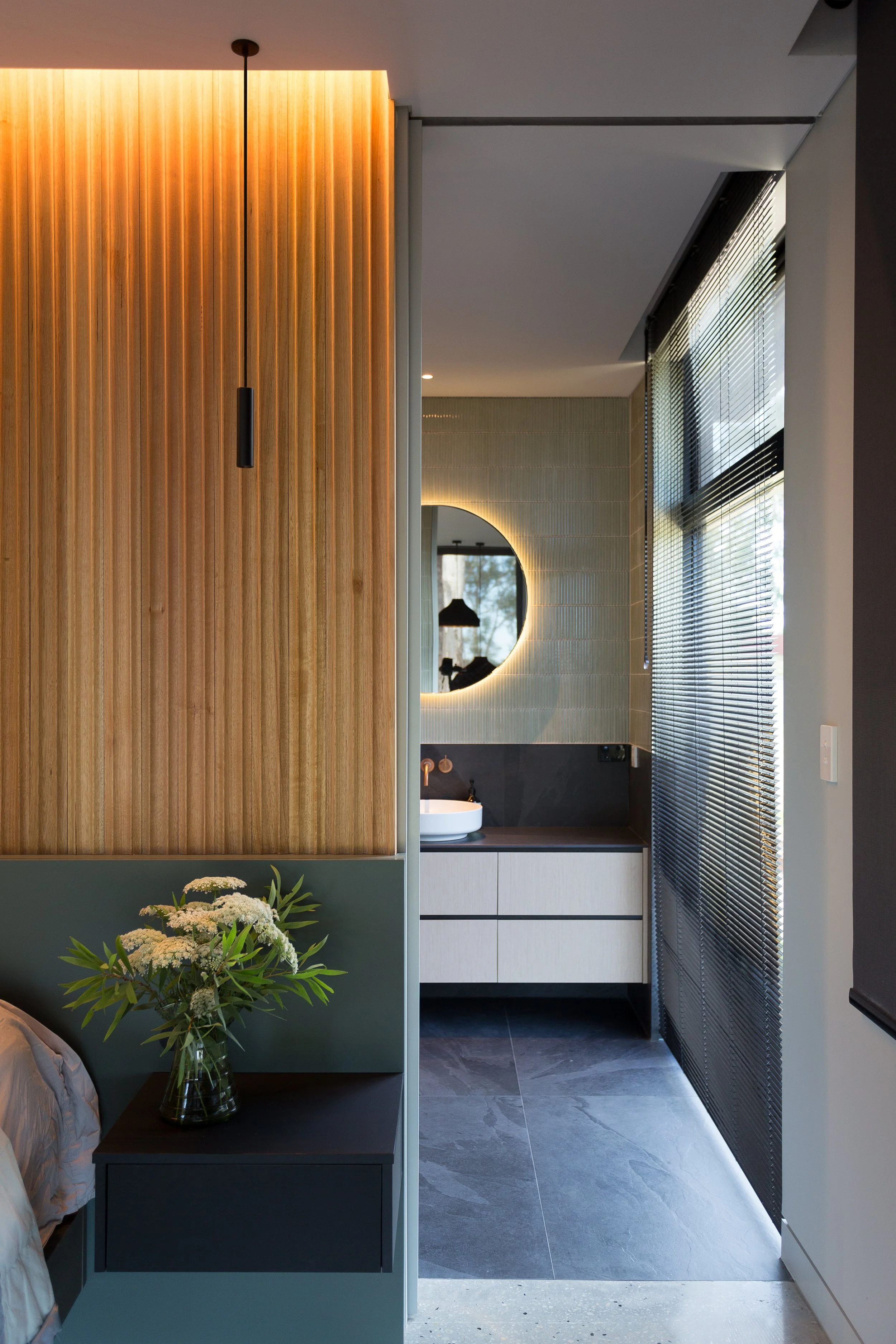 Modern interior view of a bathroom with wooden and tiled walls, a round mirror with backlighting, a vessel sink, and large windows with blinds.