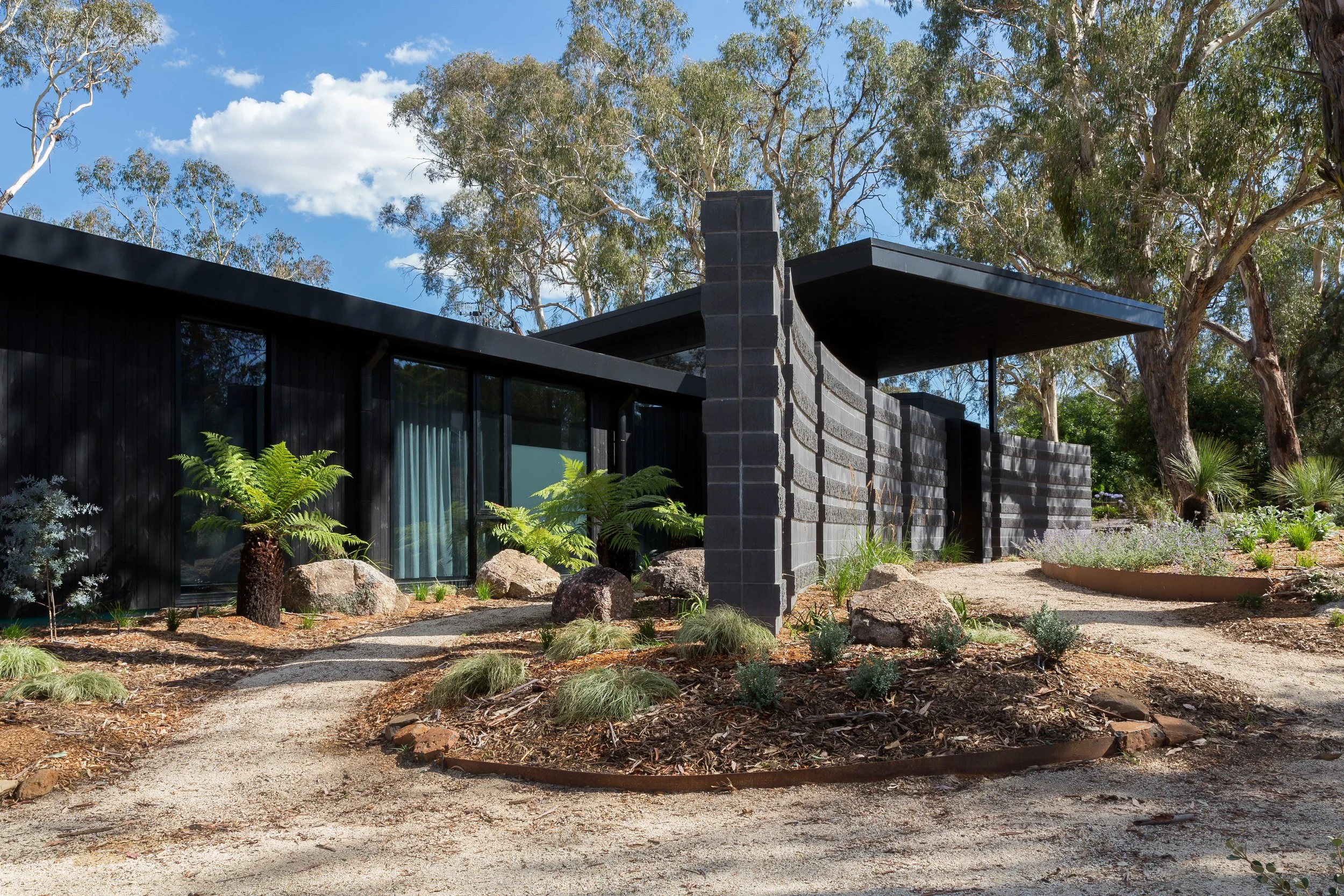 Modern black house with large windows, surrounded by trees and plants, with a gravel pathway leading to the entrance, under a blue sky with scattered clouds.