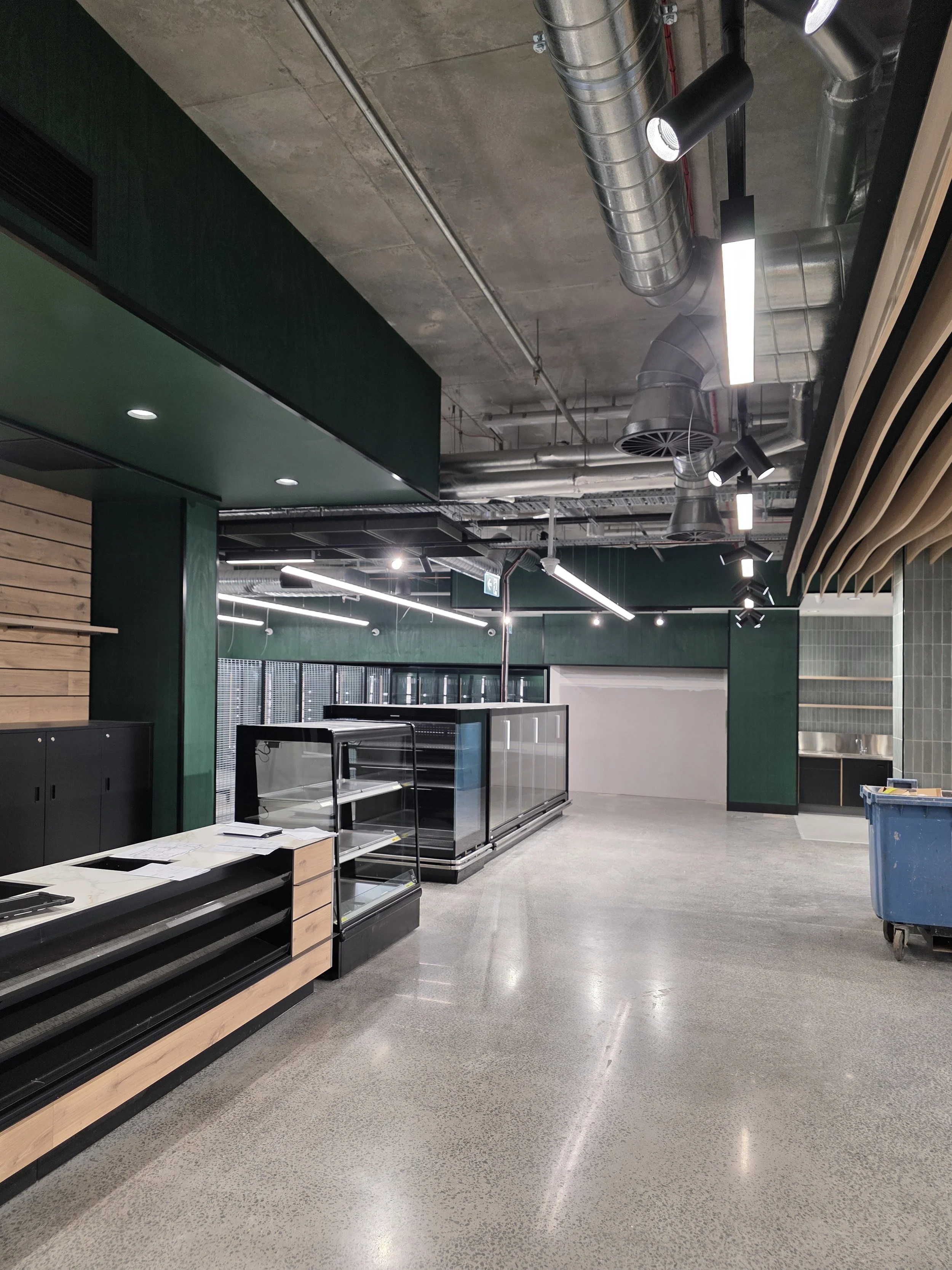 Empty retail store space with black shelves, glass display cases, and a blue trash bin. The ceiling has exposed ductwork and modern lighting. The floor is polished concrete.