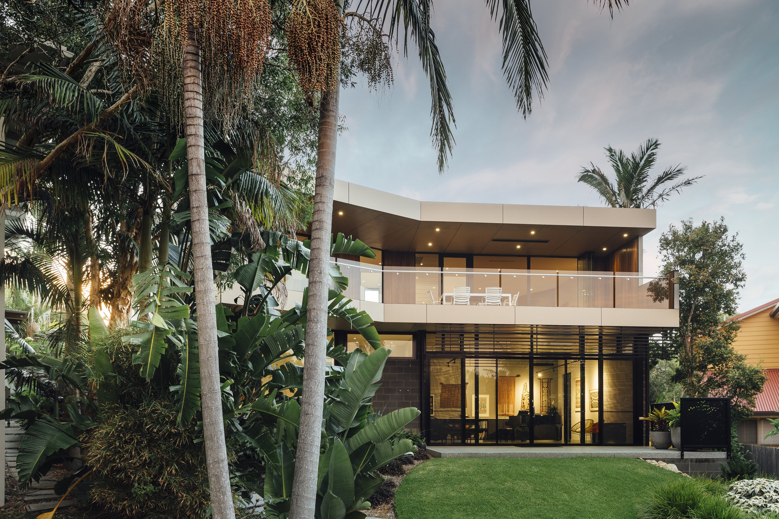 Modern two-story house with large glass windows and balconies, surrounded by tall palm trees and lush greenery, during the evening.