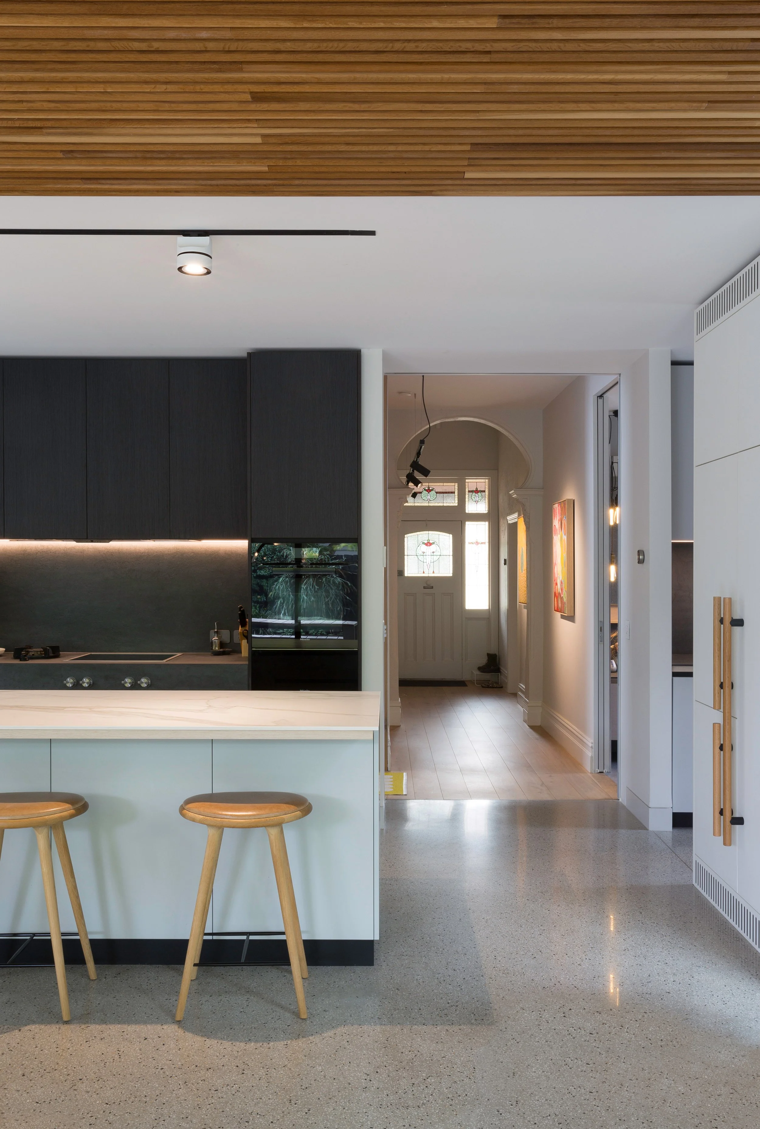 Modern kitchen with a white island, two wooden stools, black cabinets, and a hallway with a front door with stained glass windows.